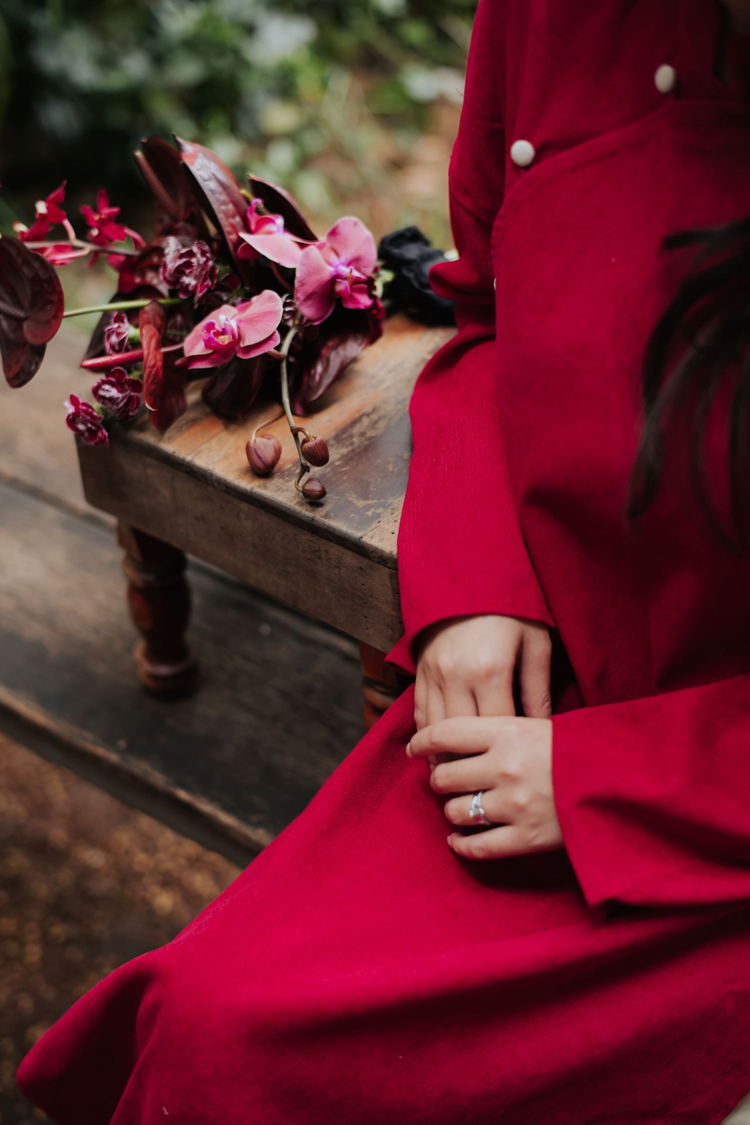 Person in red dress sitting on wooden bench with pink and burgundy floral arrangement nearby.