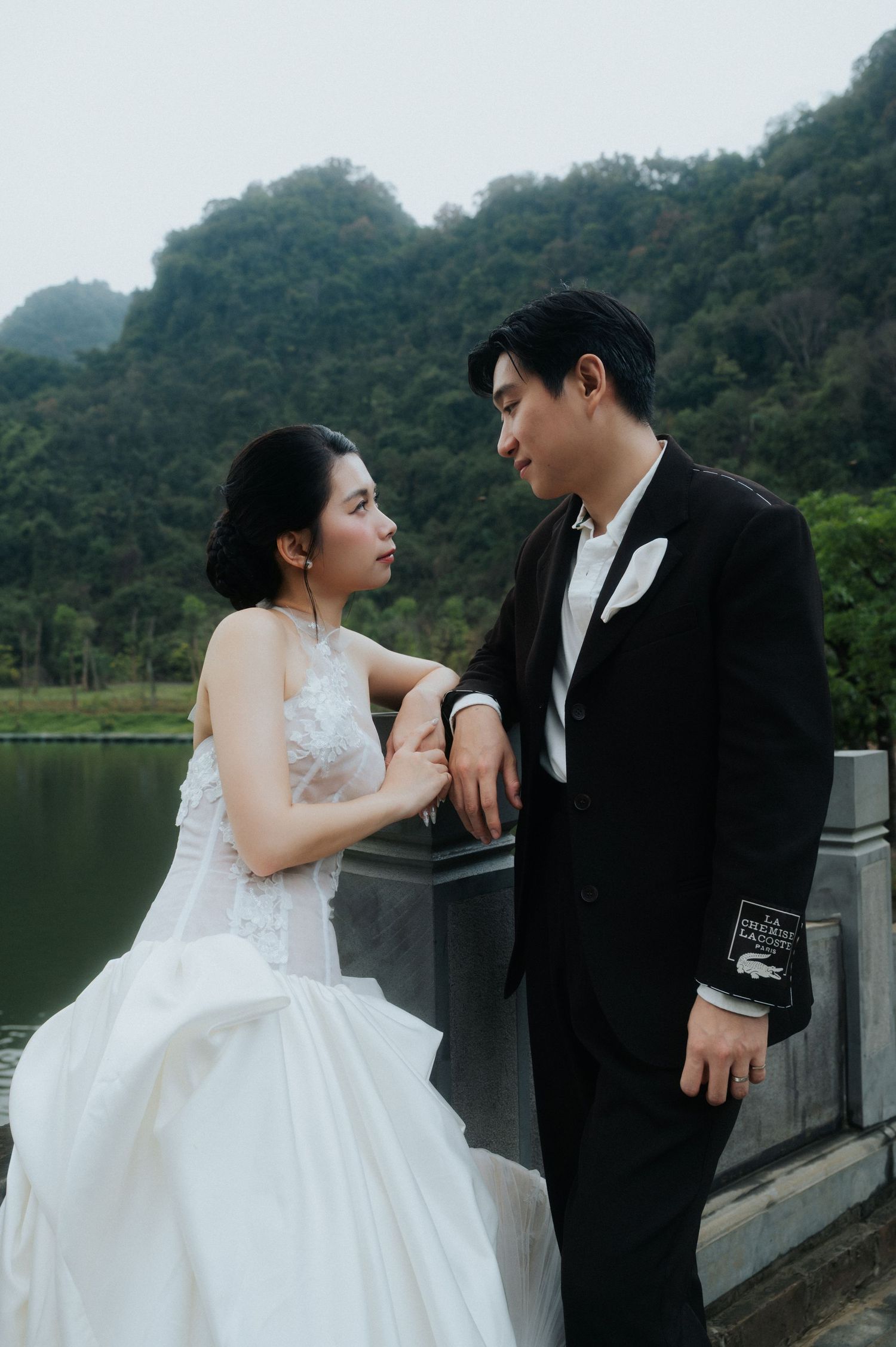 A couple in wedding attire poses romantically by a lakeside with lush green mountains in the background.