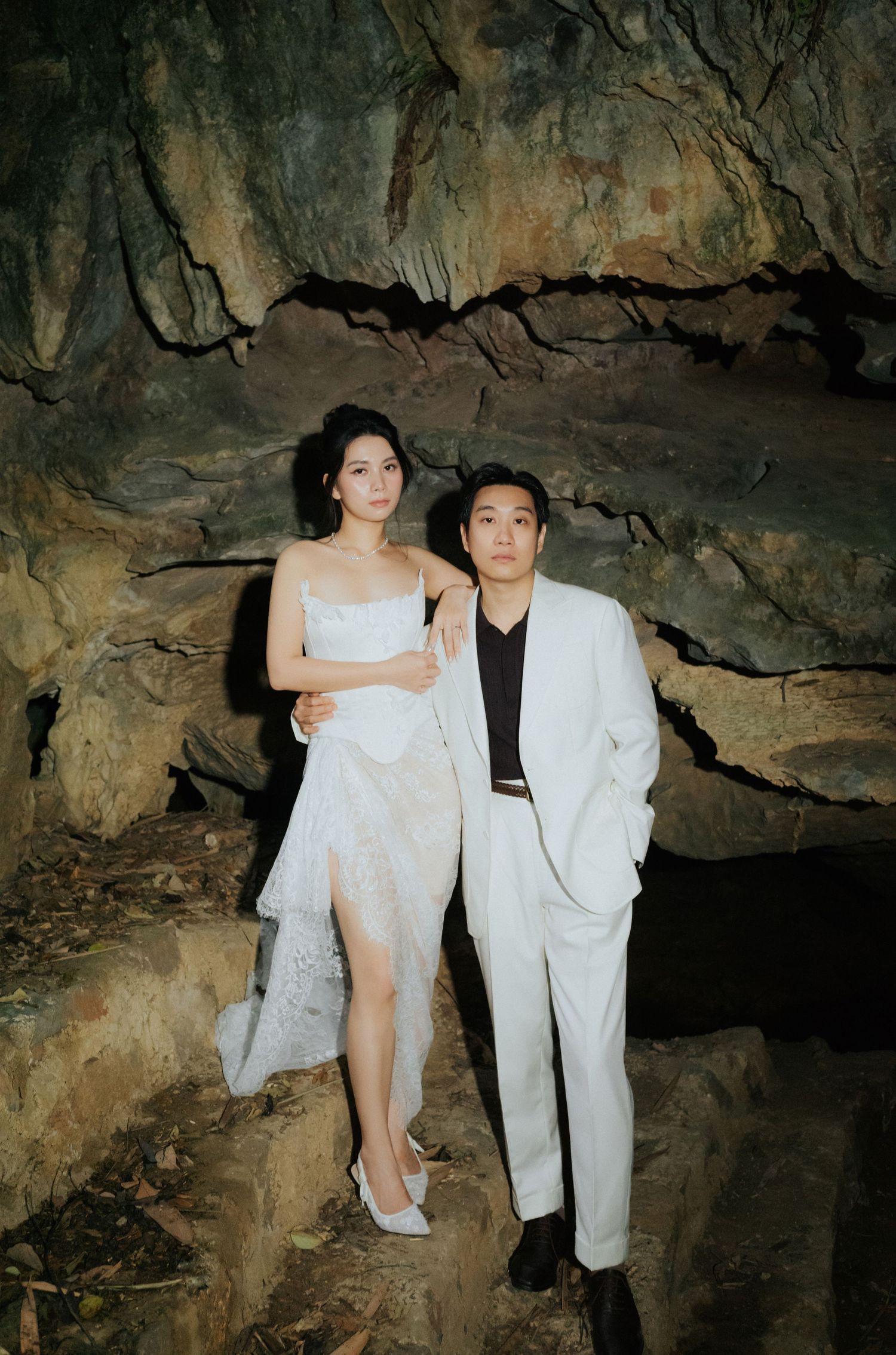 Couple in white outfits posing elegantly inside a dramatic cave with rocky walls.