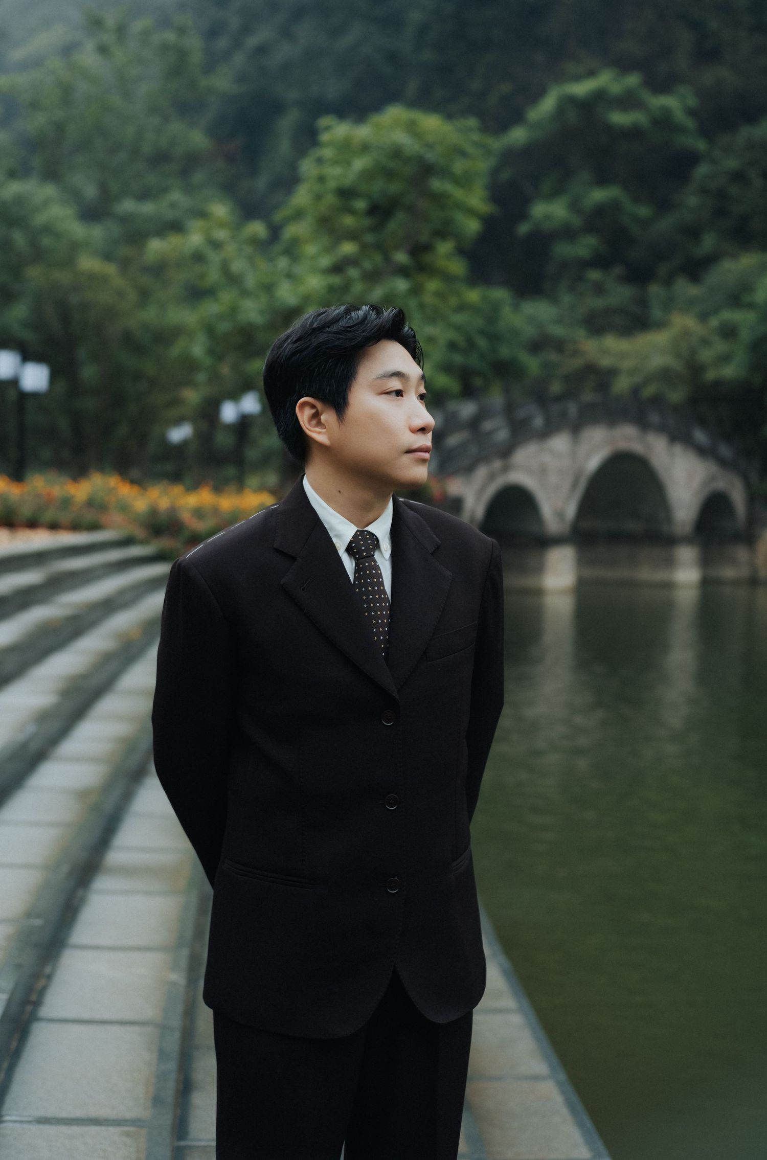 A young Asian man in a black suit and tie stands by a tranquil pond with a stone bridge and lush trees behind him.