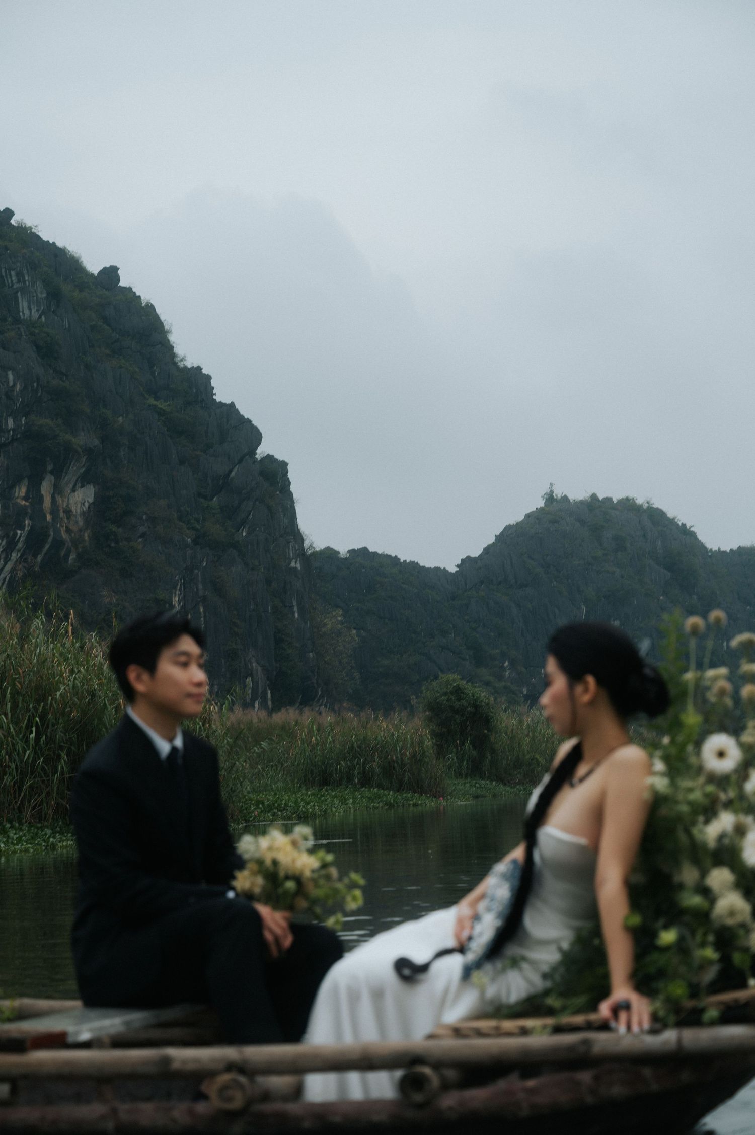 Couple in wedding attire sitting on a boat surrounded by flowers with misty karst mountains in the background.