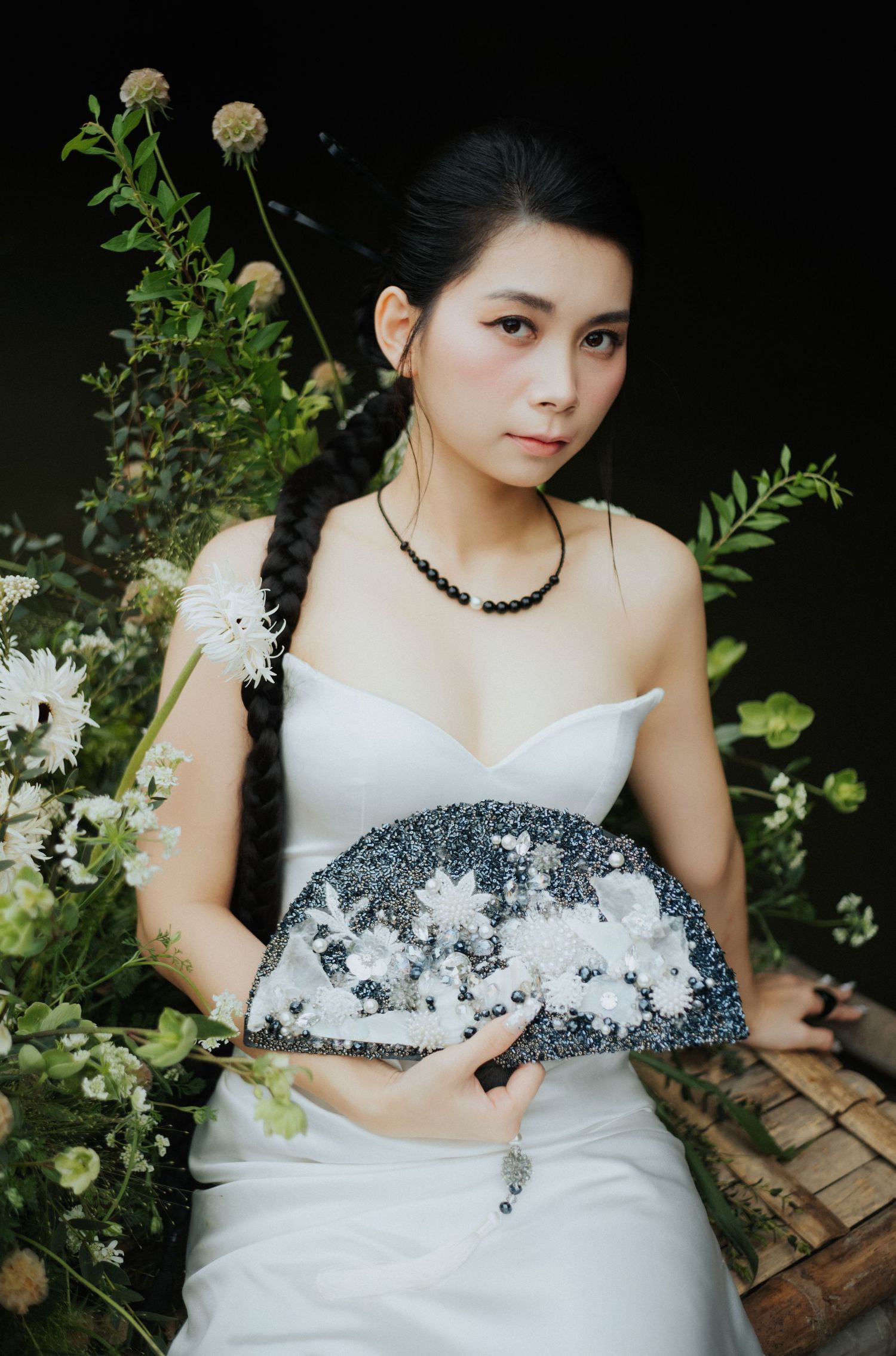 Young Asian woman in white strapless dress holding decorative fan, surrounded by white flowers and green foliage.
