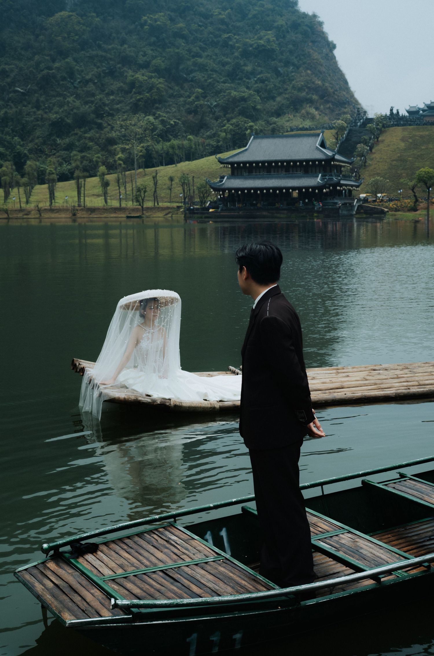 Man in black suit stands on wooden boat by serene lake with traditional Asian temple and green hills backdrop.