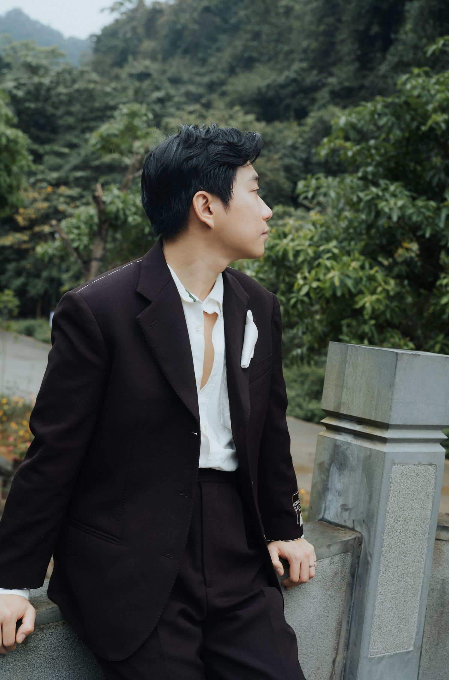 Young Asian man in dark suit with white pocket square leans on stone railing, gazing sideways amid lush green trees.