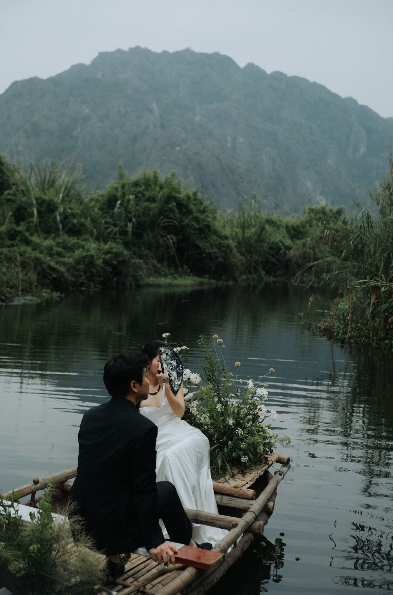 Couple in wedding attire sitting on a bamboo boat on a serene river with lush mountains in the background.