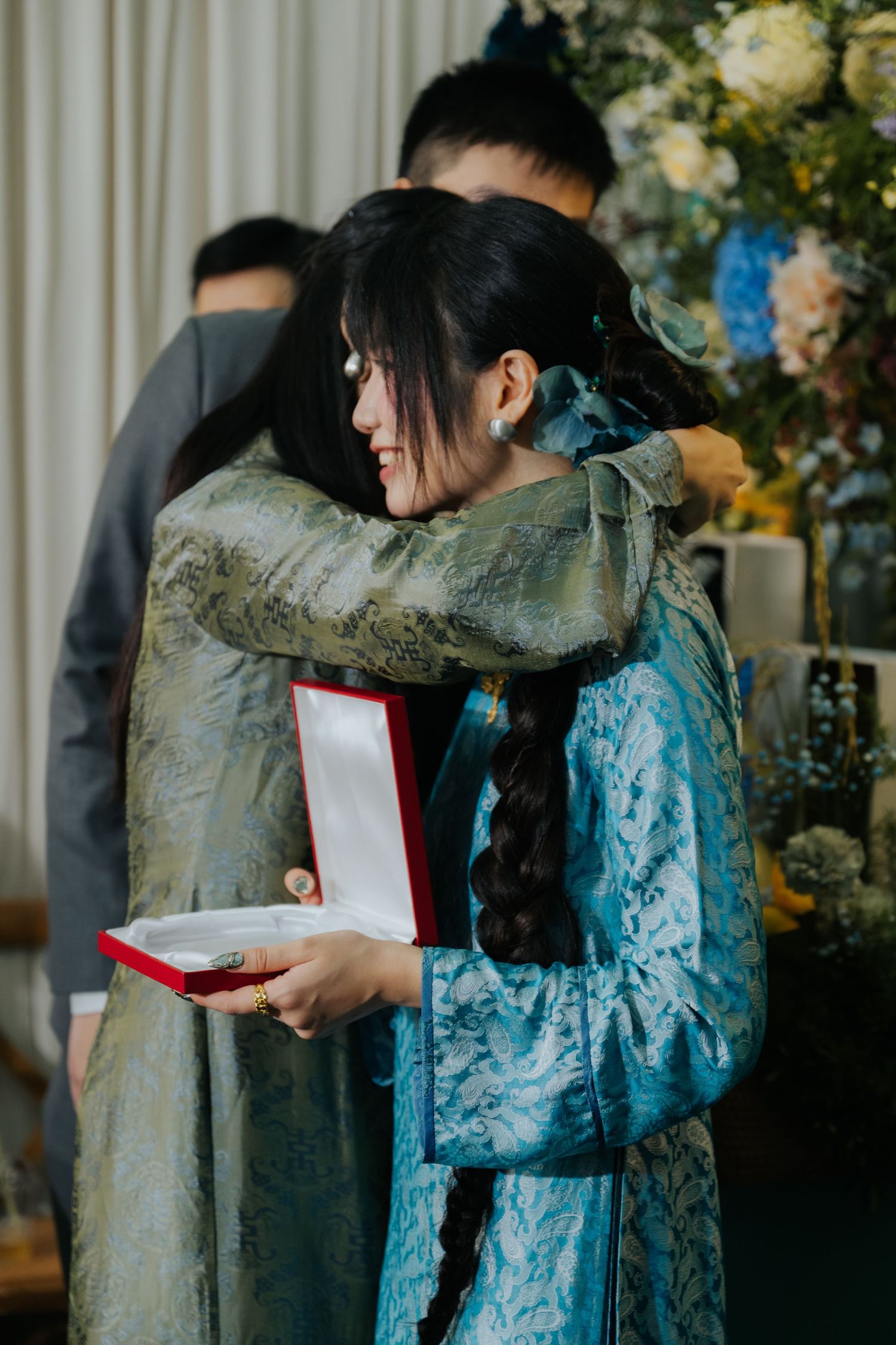 Two women in traditional Vietnamese ao dai embrace warmly during a wedding ceremony while holding a red jewelry gift box.