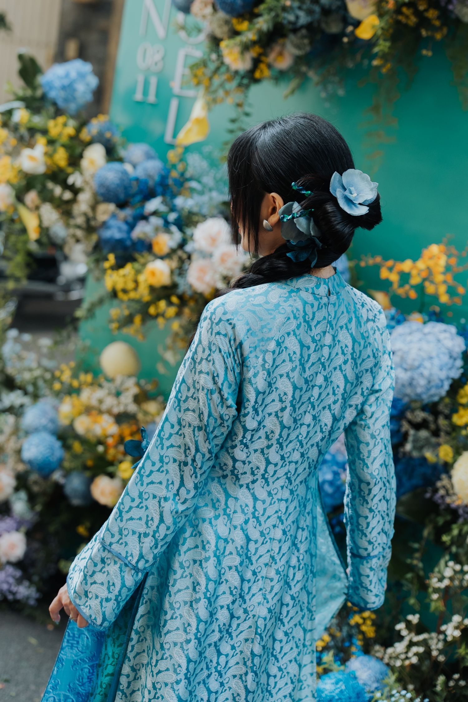 Woman in blue floral lace dress with flower hair accessory stands before colorful hydrangea floral backdrop.