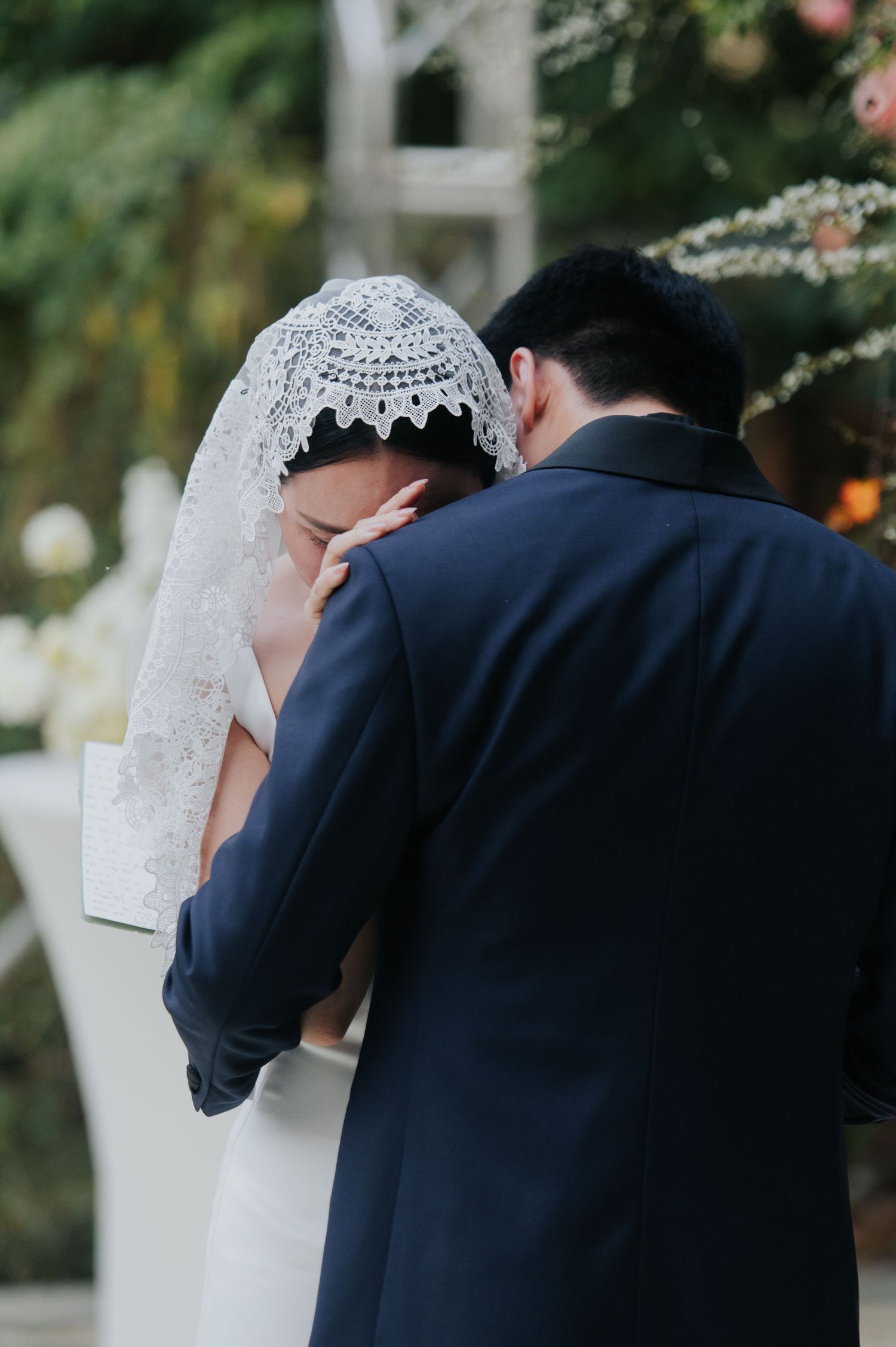 A bride in a lace veil leans on her groom in a navy suit during an emotional outdoor wedding ceremony.