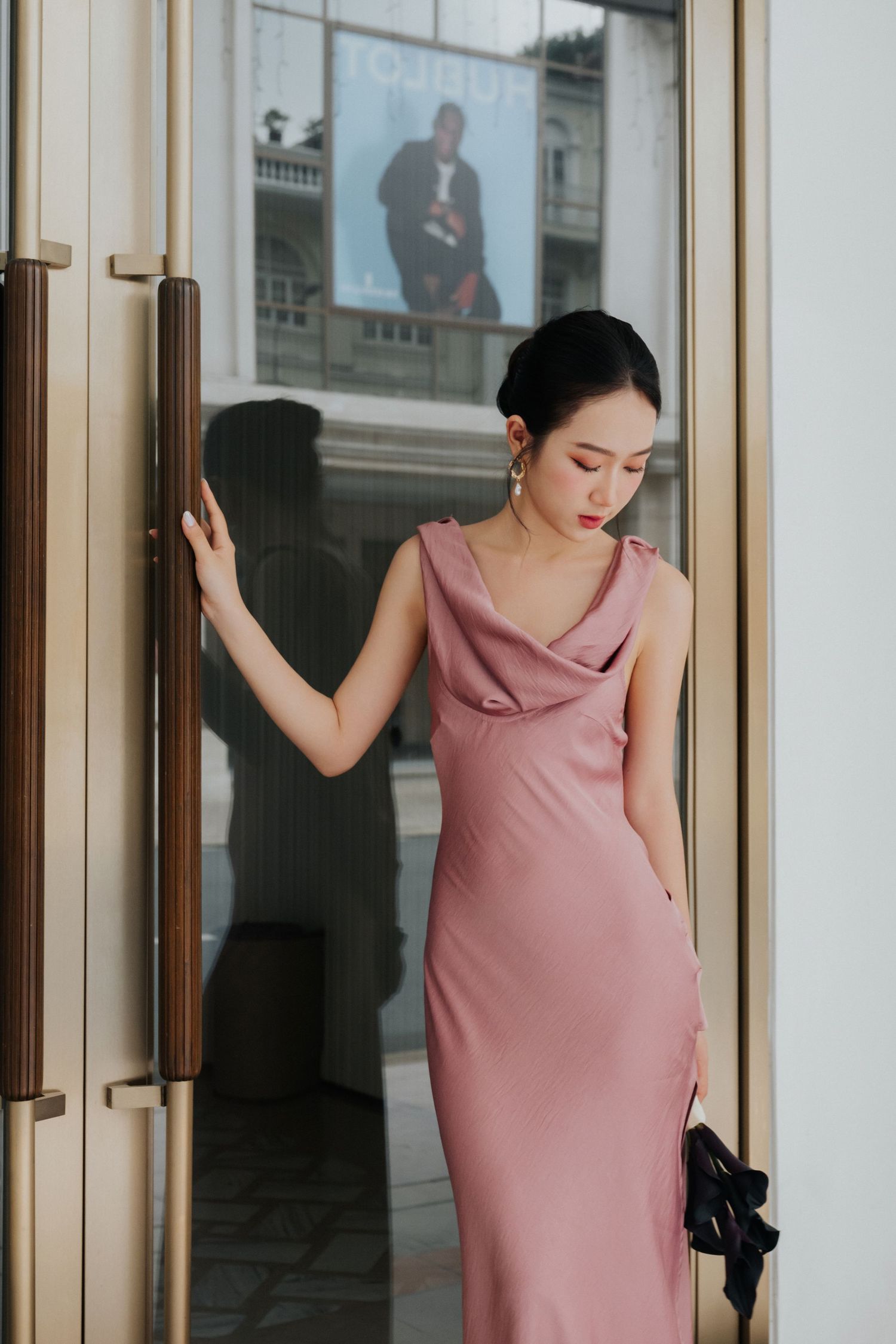 Woman in elegant pink satin slip dress poses gracefully by golden glass door of upscale building.