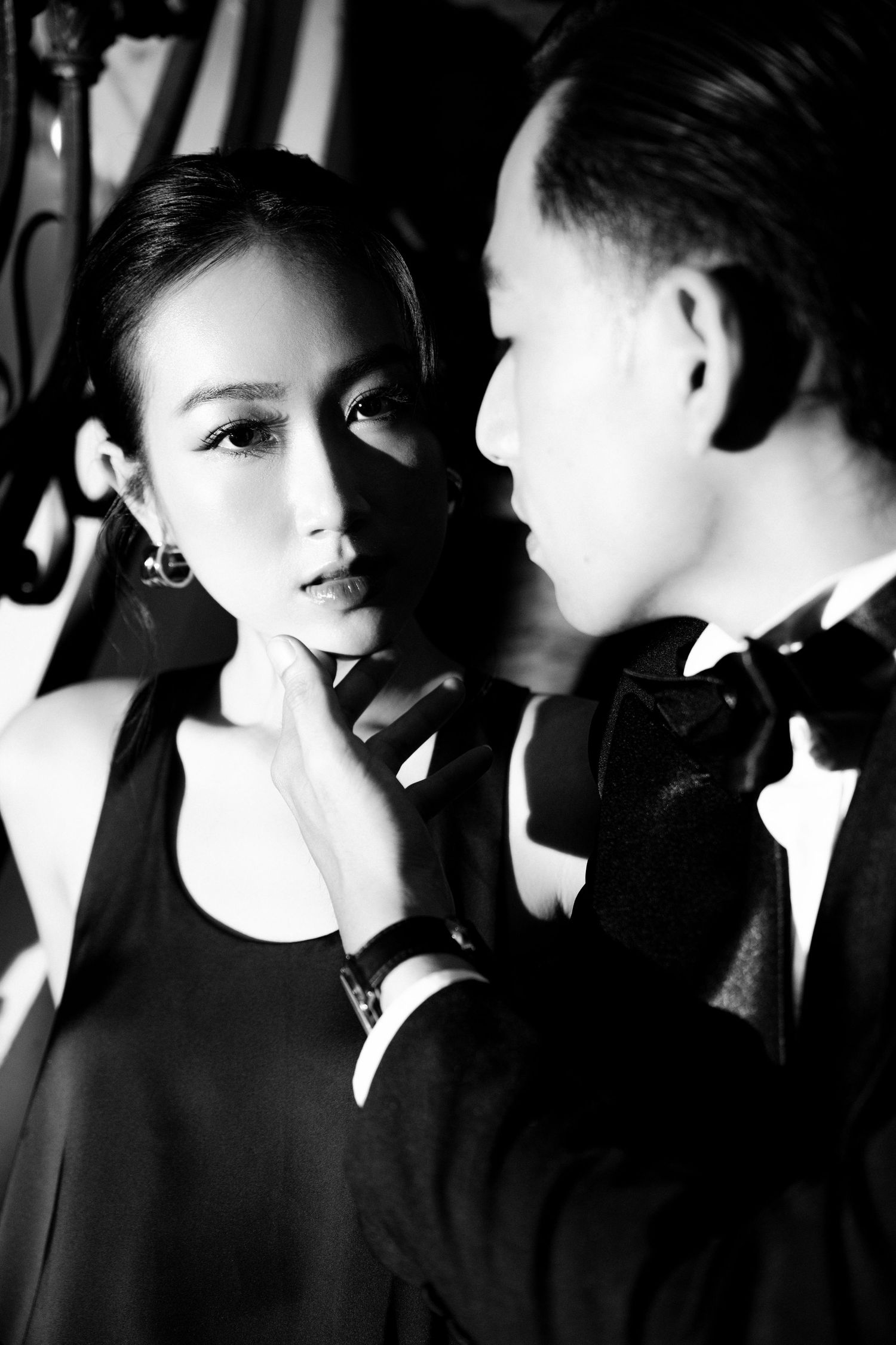 Black and white photo of an elegant couple, man in tuxedo gently touching womans chin in dramatic lighting.