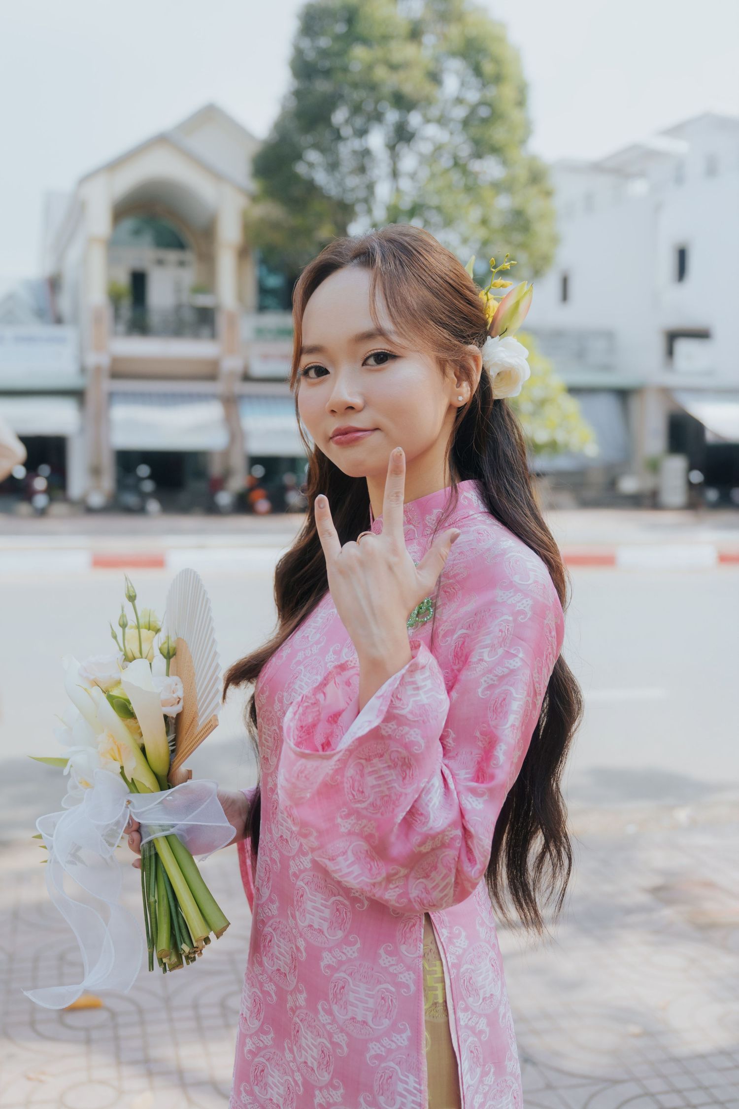 Young woman in pink floral ao dai holding white flowers, making peace sign, with flower hair accessory outdoors.