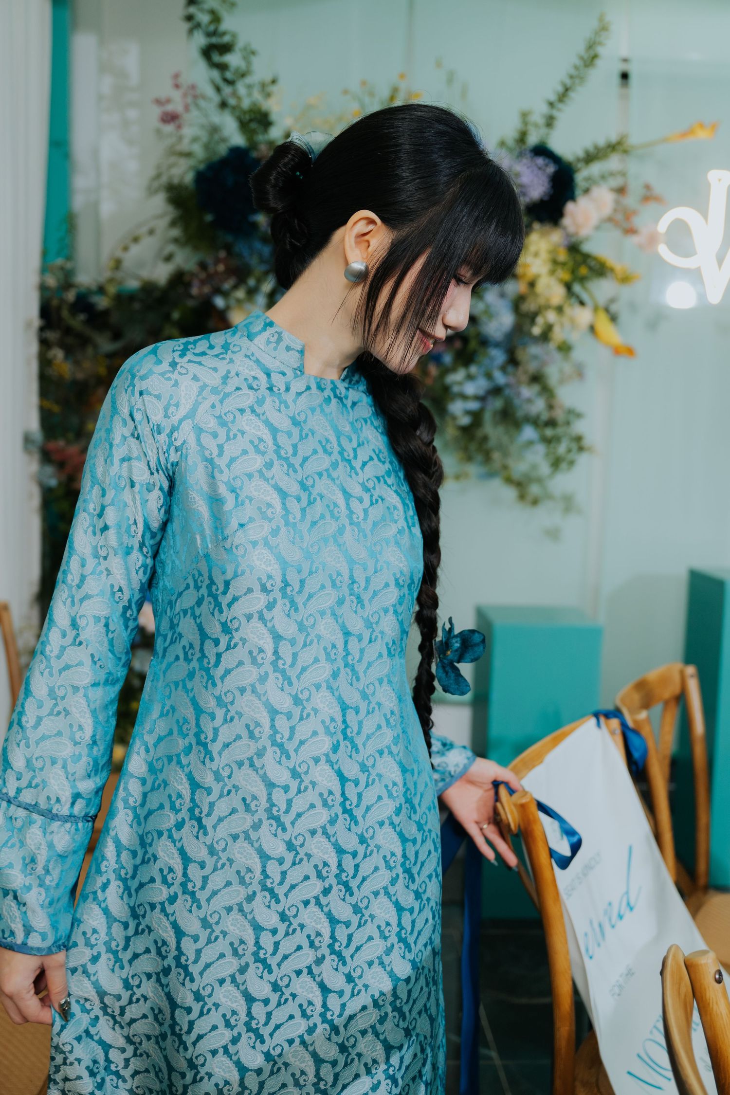 Young woman in a light blue lace ao dai dress with a braided hairstyle, standing gracefully in an elegant interior.