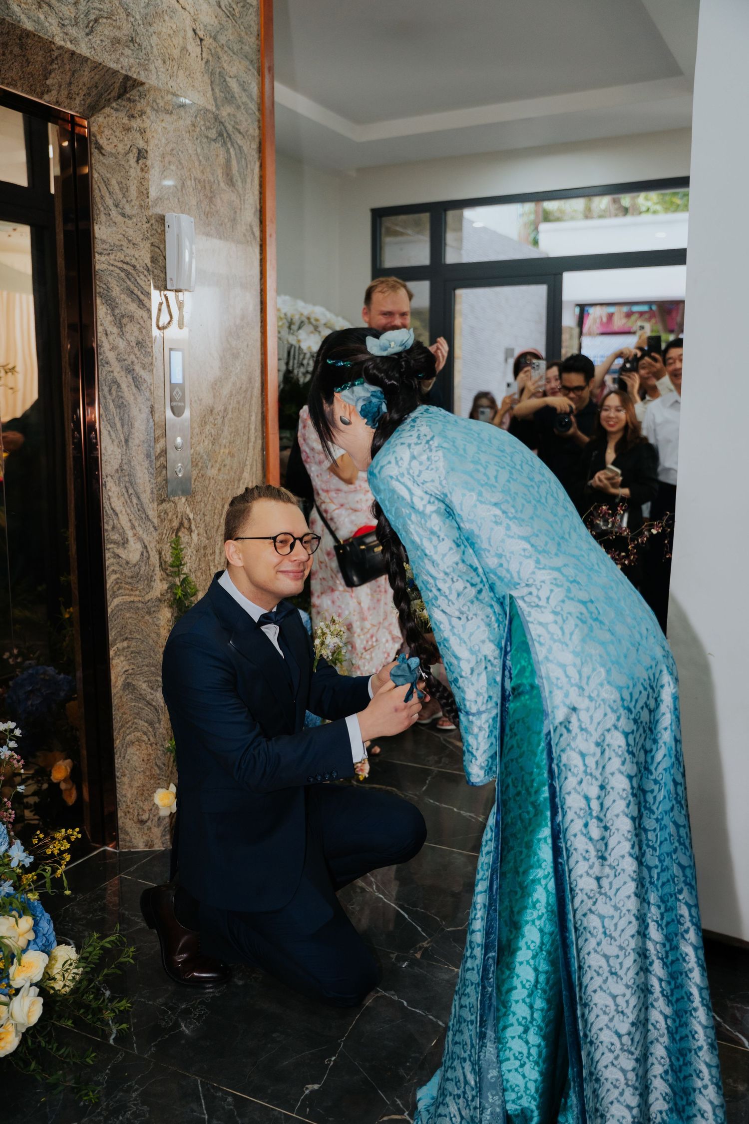 Groom kneeling before a woman in blue hijab and lace dress during a traditional wedding ceremony.