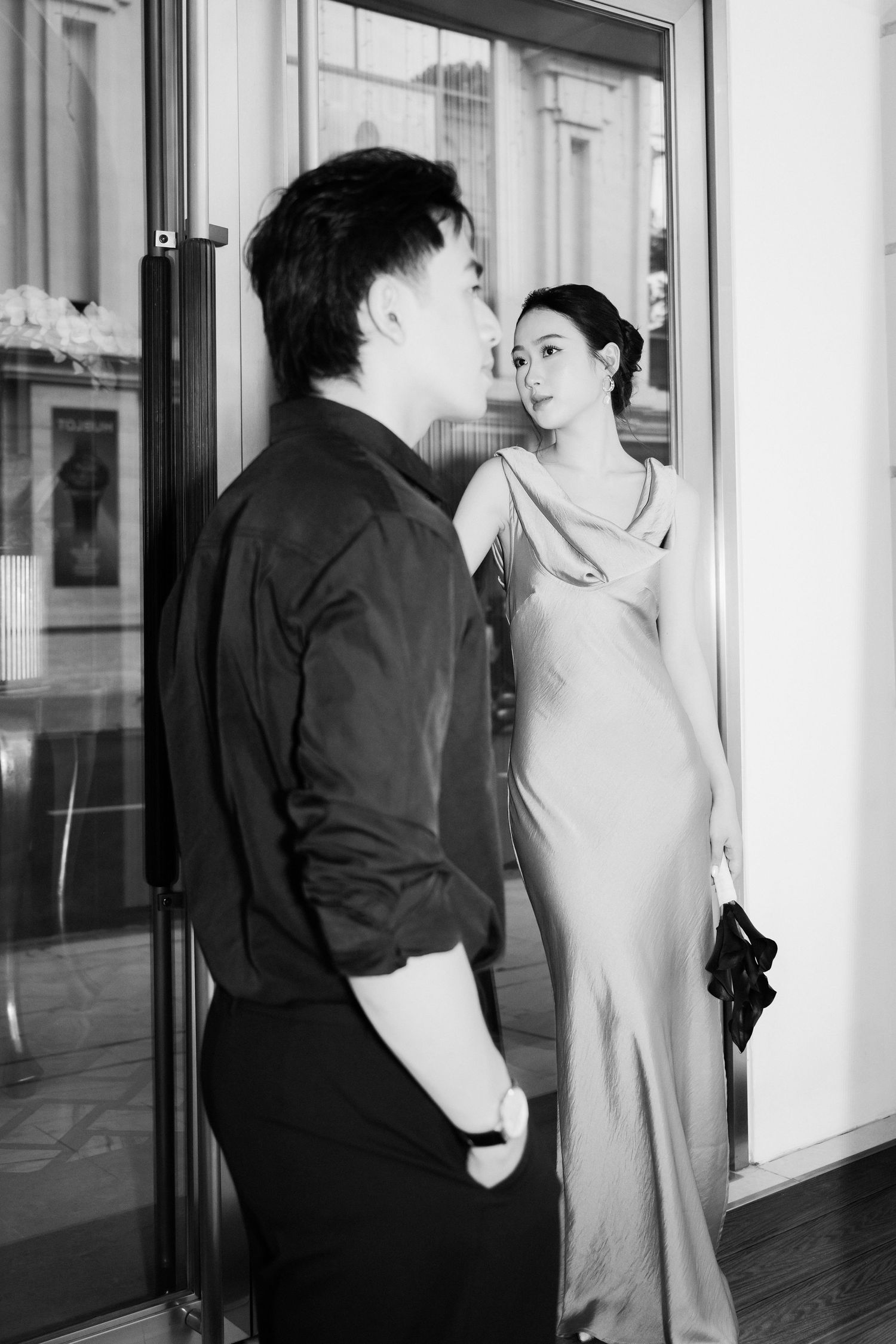 Black and white photo of a man in dark attire and a woman in an elegant slip dress exchanging glances near a glass door.