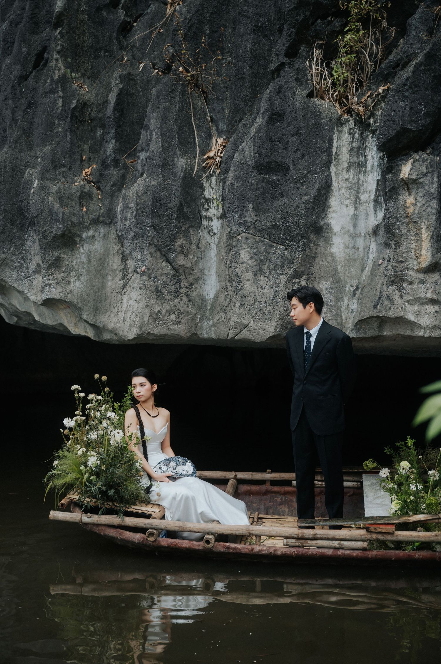 Couple in wedding attire on a bamboo boat with flowers, set against dramatic limestone cliffs over calm water.