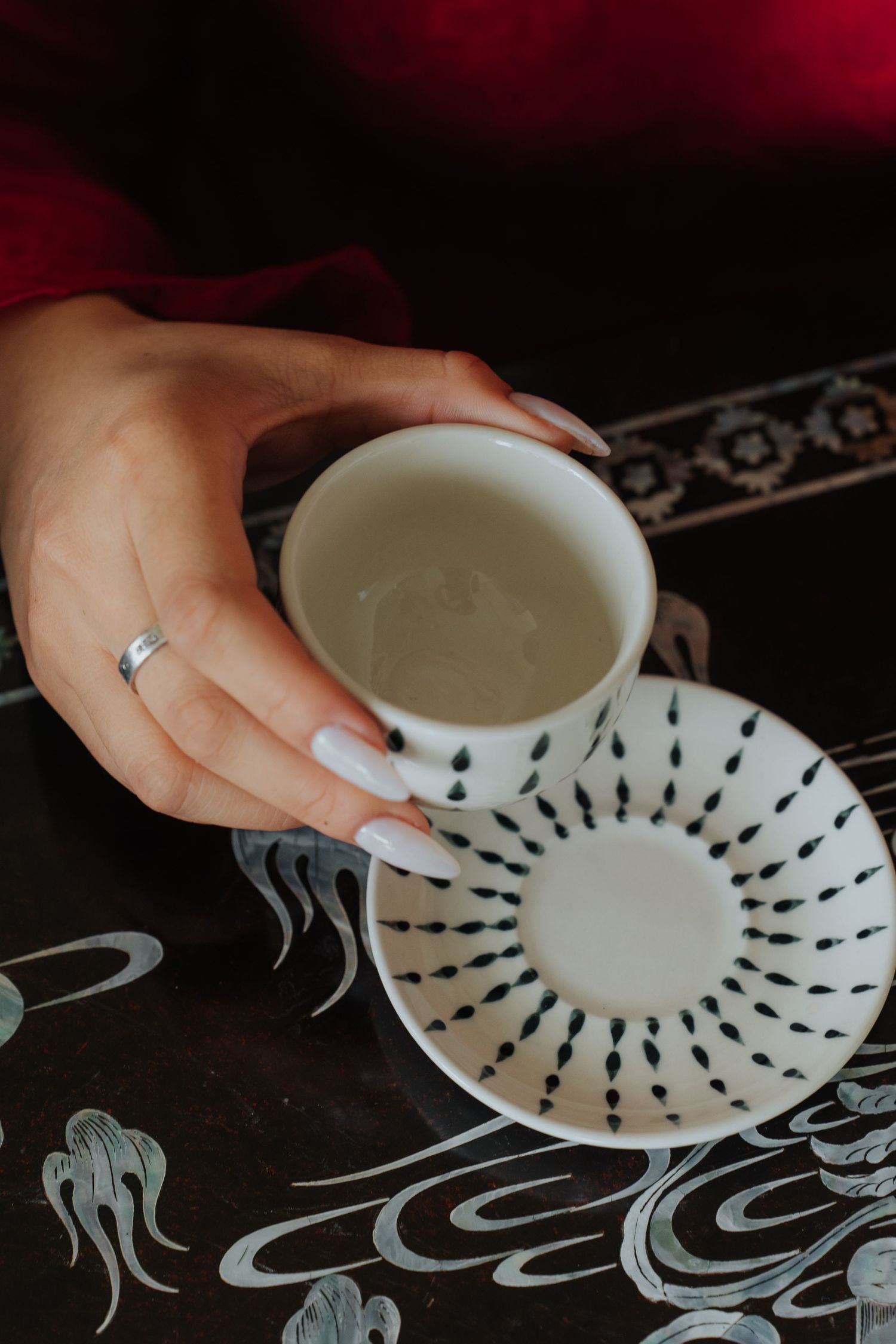 A hand with white nails and a silver ring holds a white ceramic cup with black dot pattern over a matching saucer.