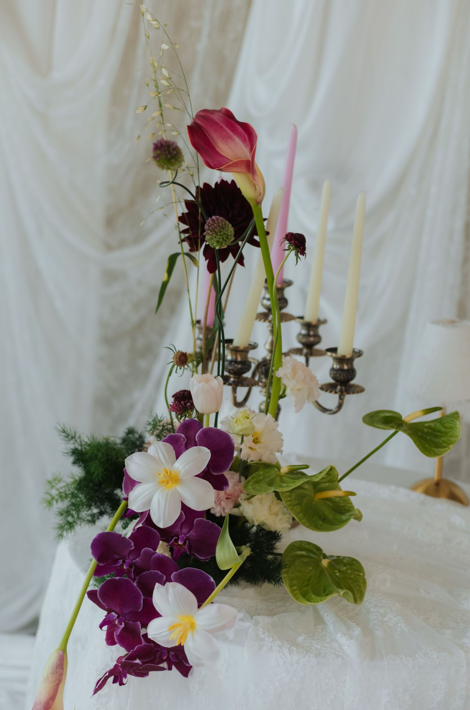 Elegant floral arrangement with purple orchids, white flowers, and a candelabra on a white lace table.