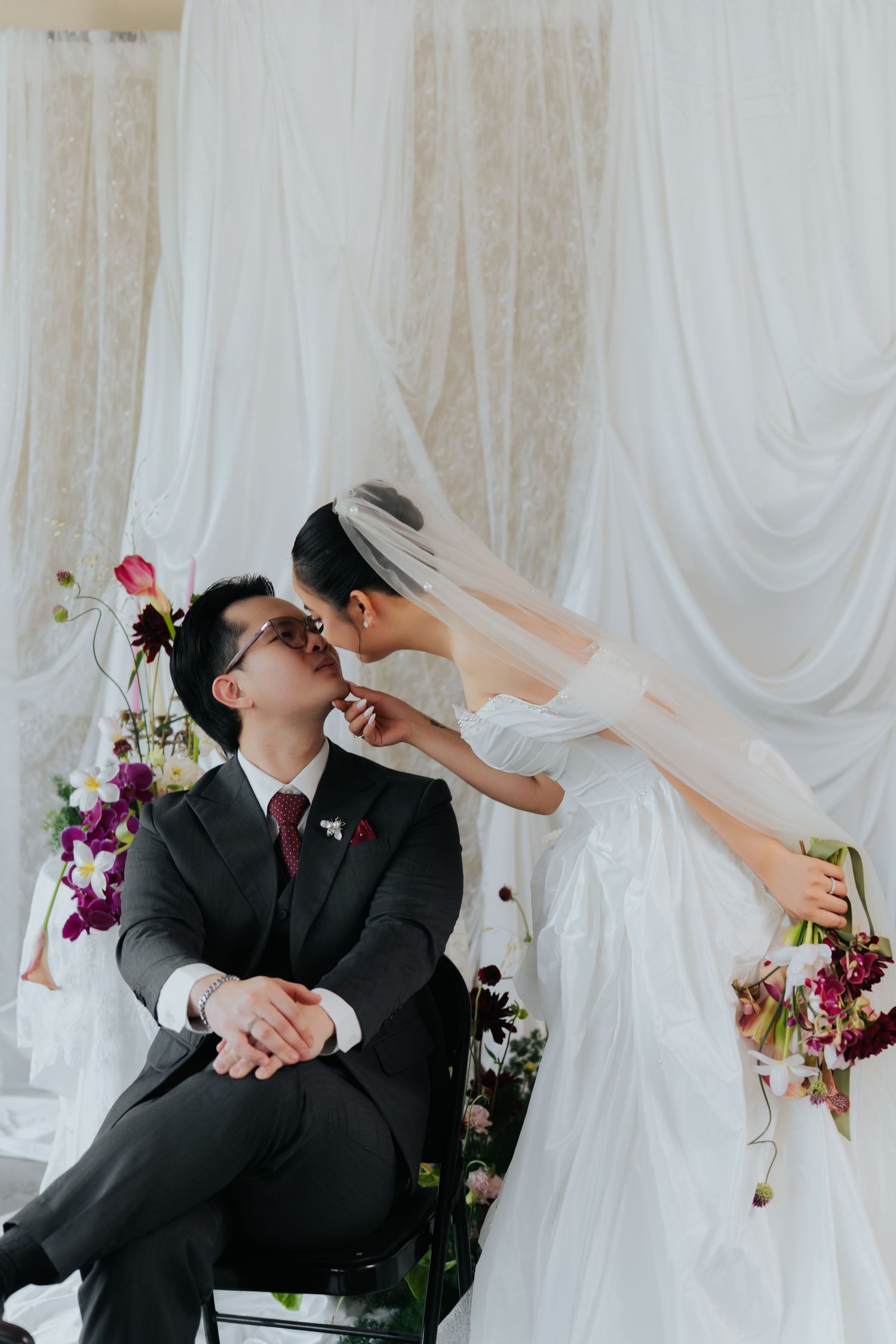 Bride in white gown and veil leans to kiss groom in dark suit, seated before elegant white draped backdrop with floral arrangements.