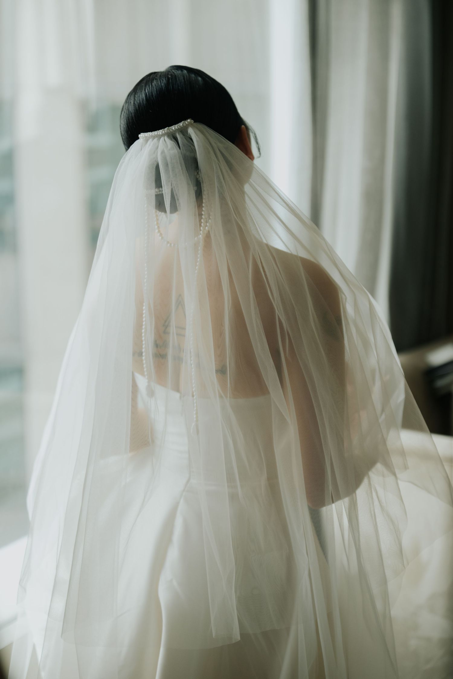 Bride in white wedding dress and long veil seen from behind, standing near window in soft natural light.