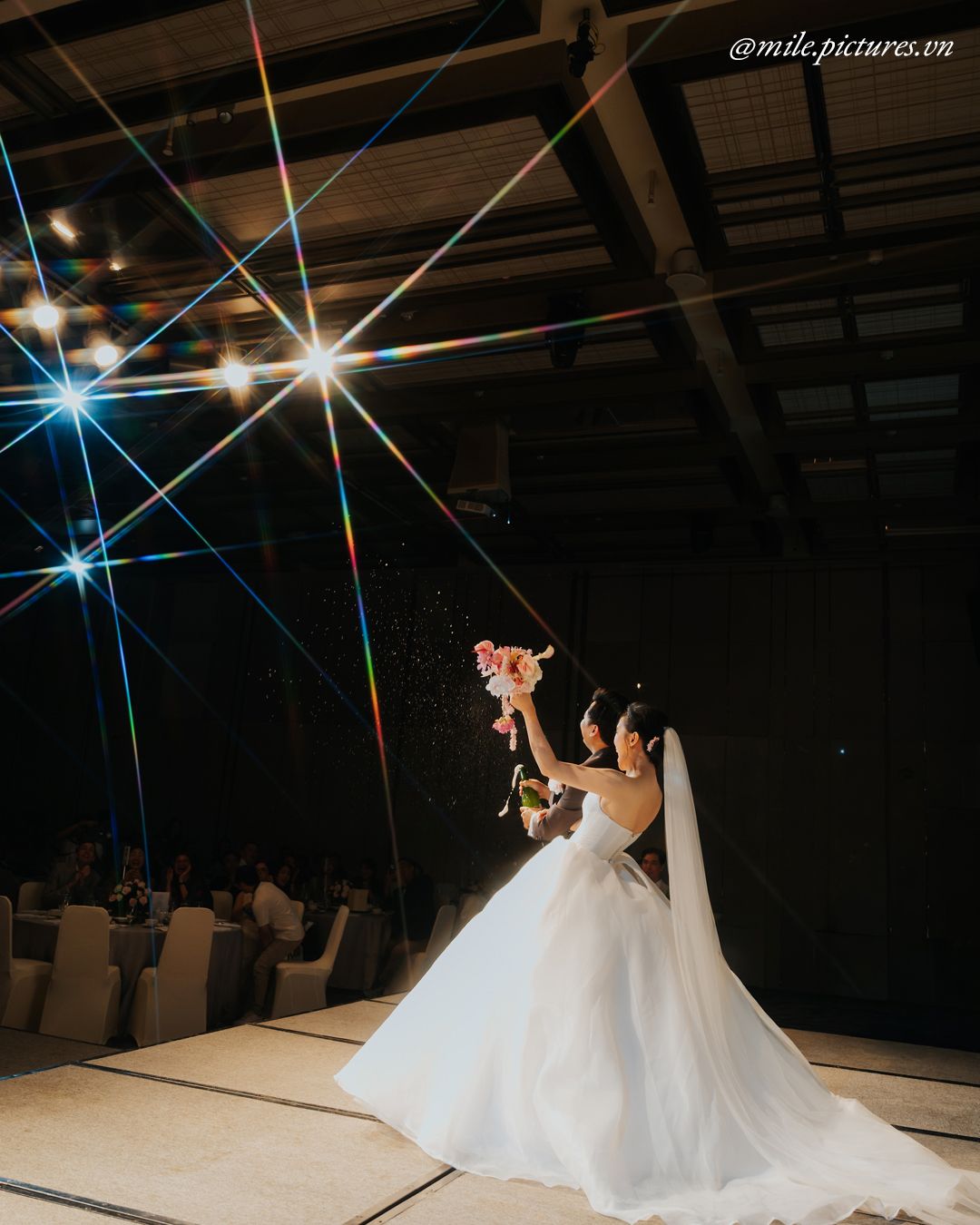 Bride in white gown holds bouquet aloft under dramatic starburst lights at elegant wedding reception.