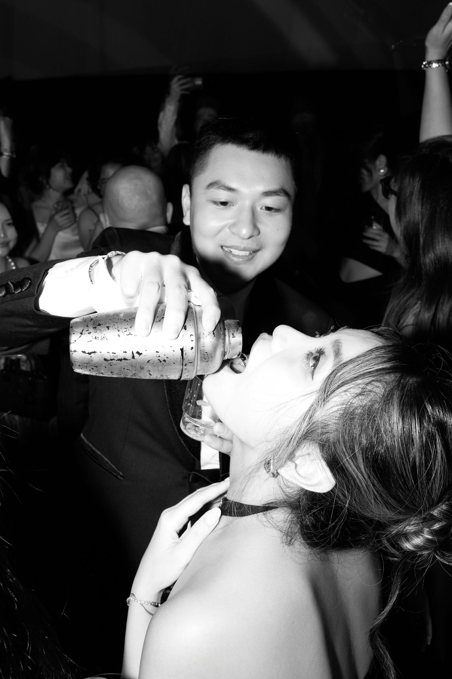 Black and white photo of a man pouring a drink into a woman's mouth at a lively party.