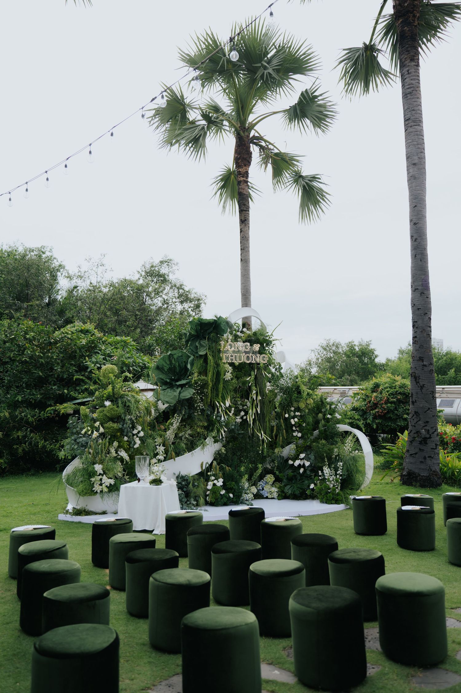 Outdoor wedding ceremony setup with green ottomans, floral arch, and palm trees on a lush lawn.