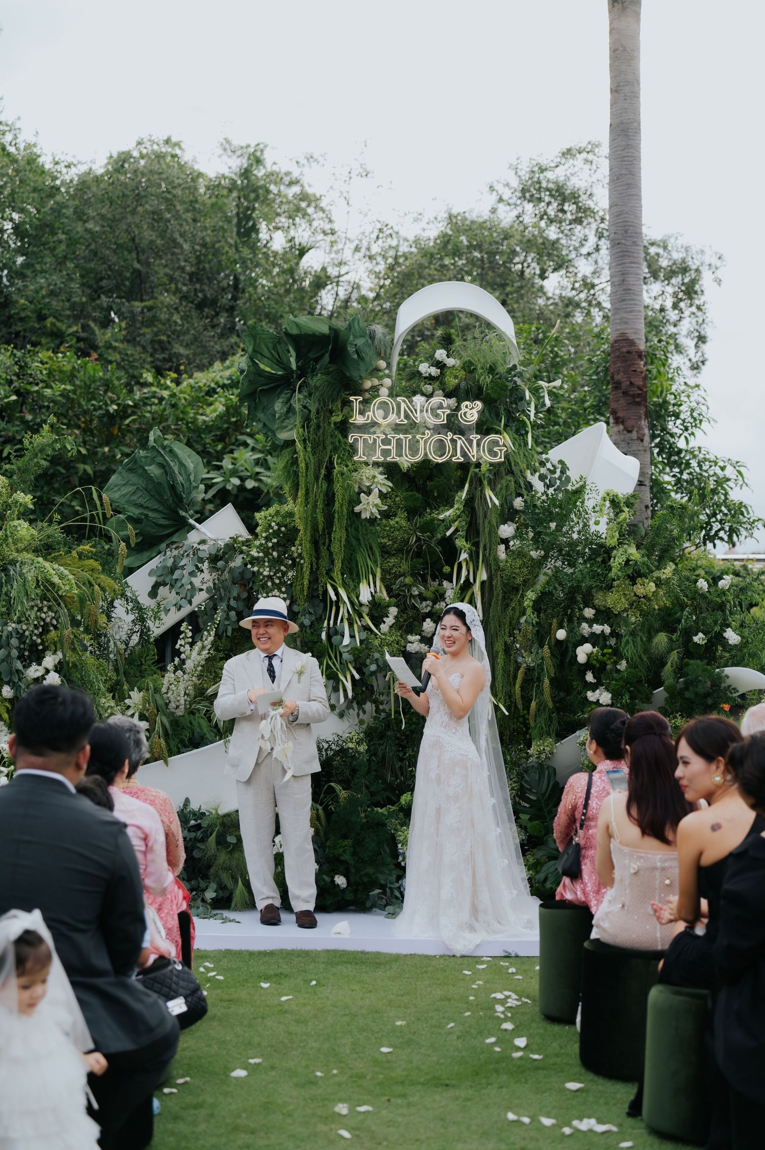 Outdoor wedding ceremony with bride in white gown and groom in cream suit at floral arch reading Long & Thuong.