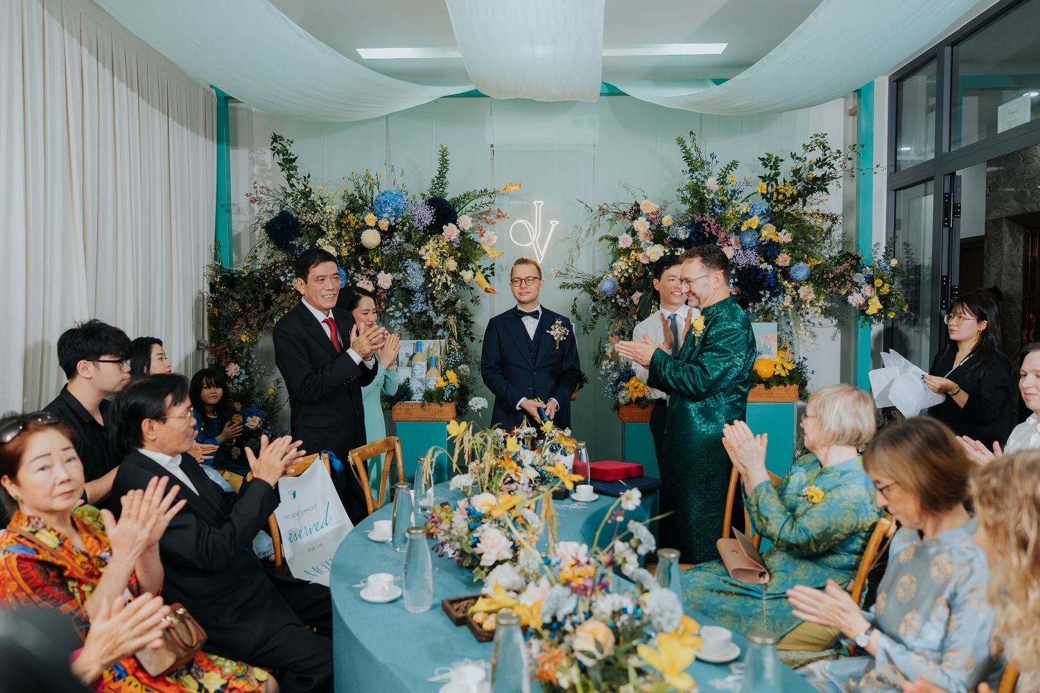 Two grooms celebrate at a elegant wedding reception with floral arrangements in blue and yellow tones, surrounded by applauding guests.