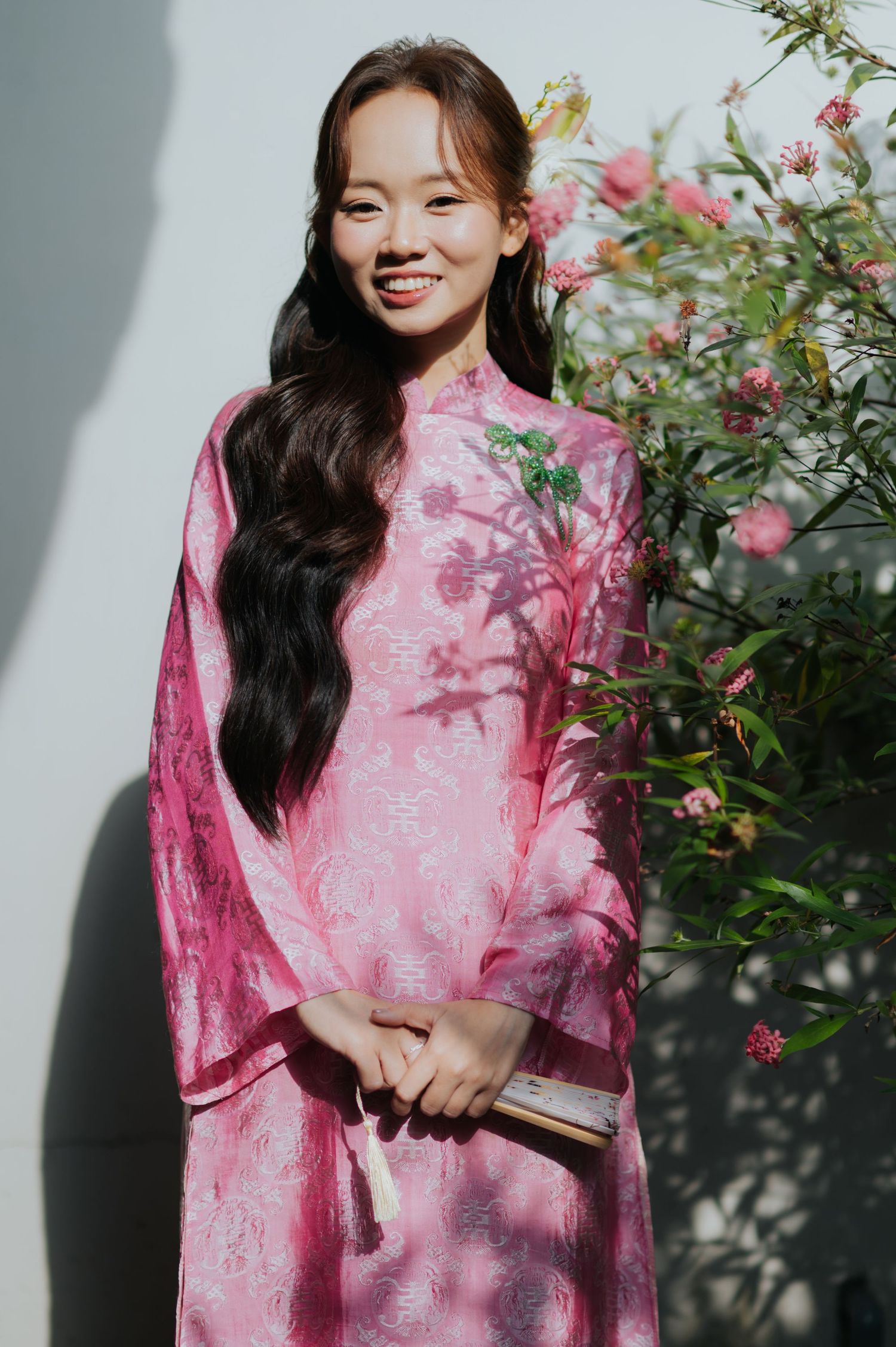Young woman smiling in a pink floral áo dài, holding a fan, standing beside blooming pink flowers outdoors.