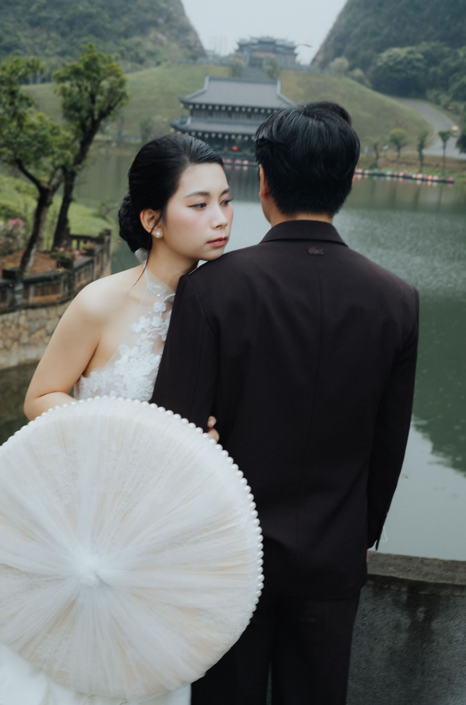 A bride in a white lace gown holds a parasol beside a groom in a black suit near a serene lake and pagoda.