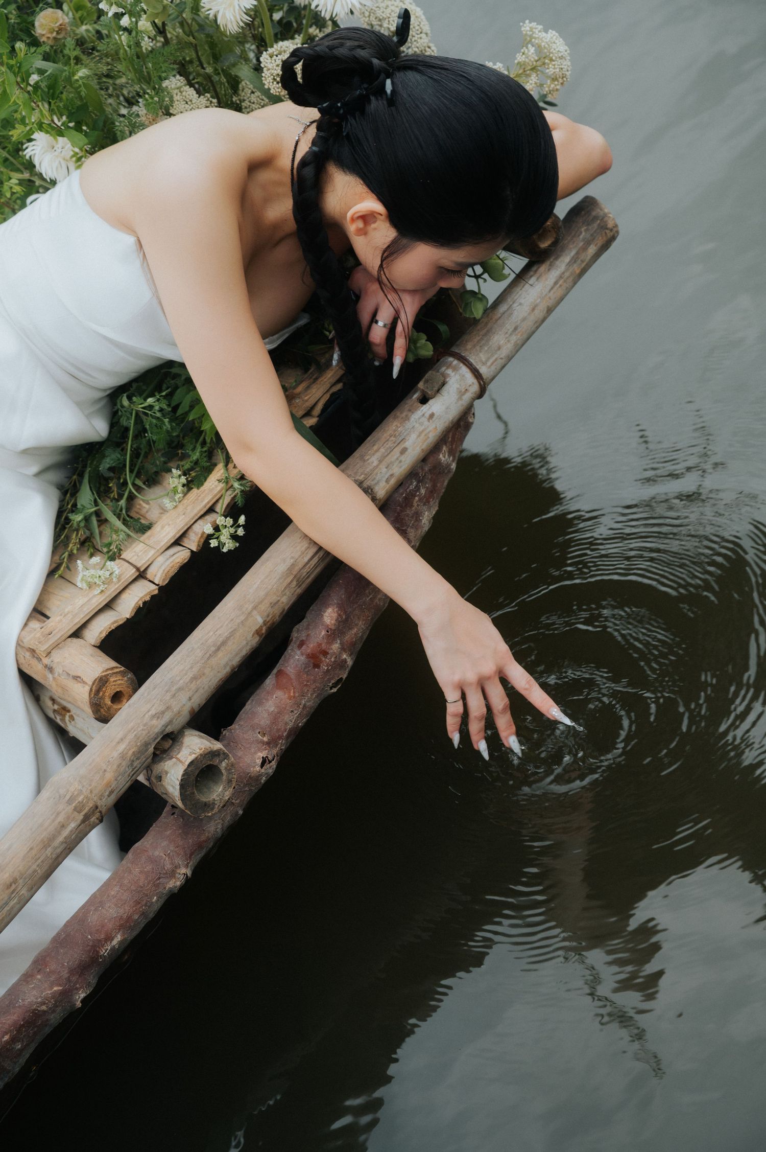 A woman in a white dress leans over a wooden boat, gently touching the dark water with her fingertips.