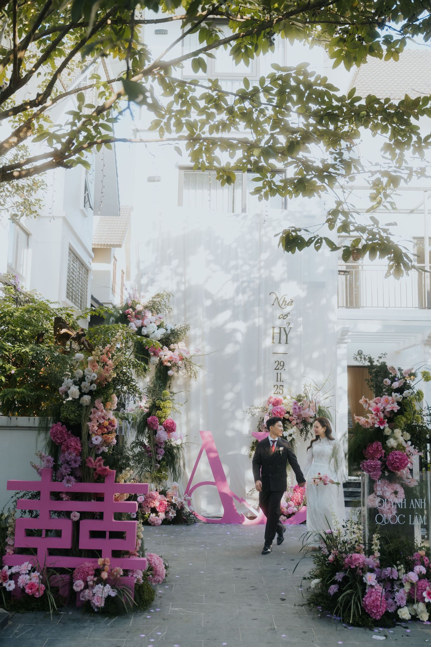 Couple walks through floral wedding arch with pink blooms and decorative letters at an outdoor ceremony.
