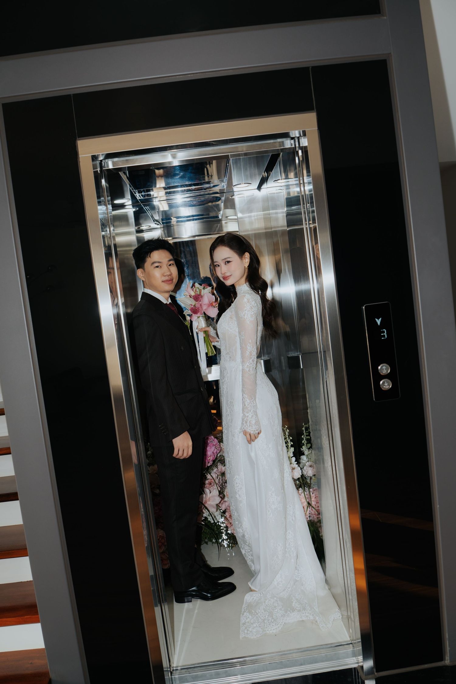 A bride in a white lace gown and groom in a black suit pose together inside a glass elevator.