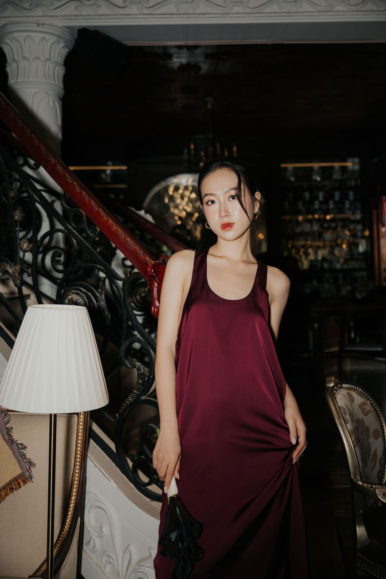 Woman in elegant burgundy satin slip dress poses on ornate staircase in luxurious dark interior setting.