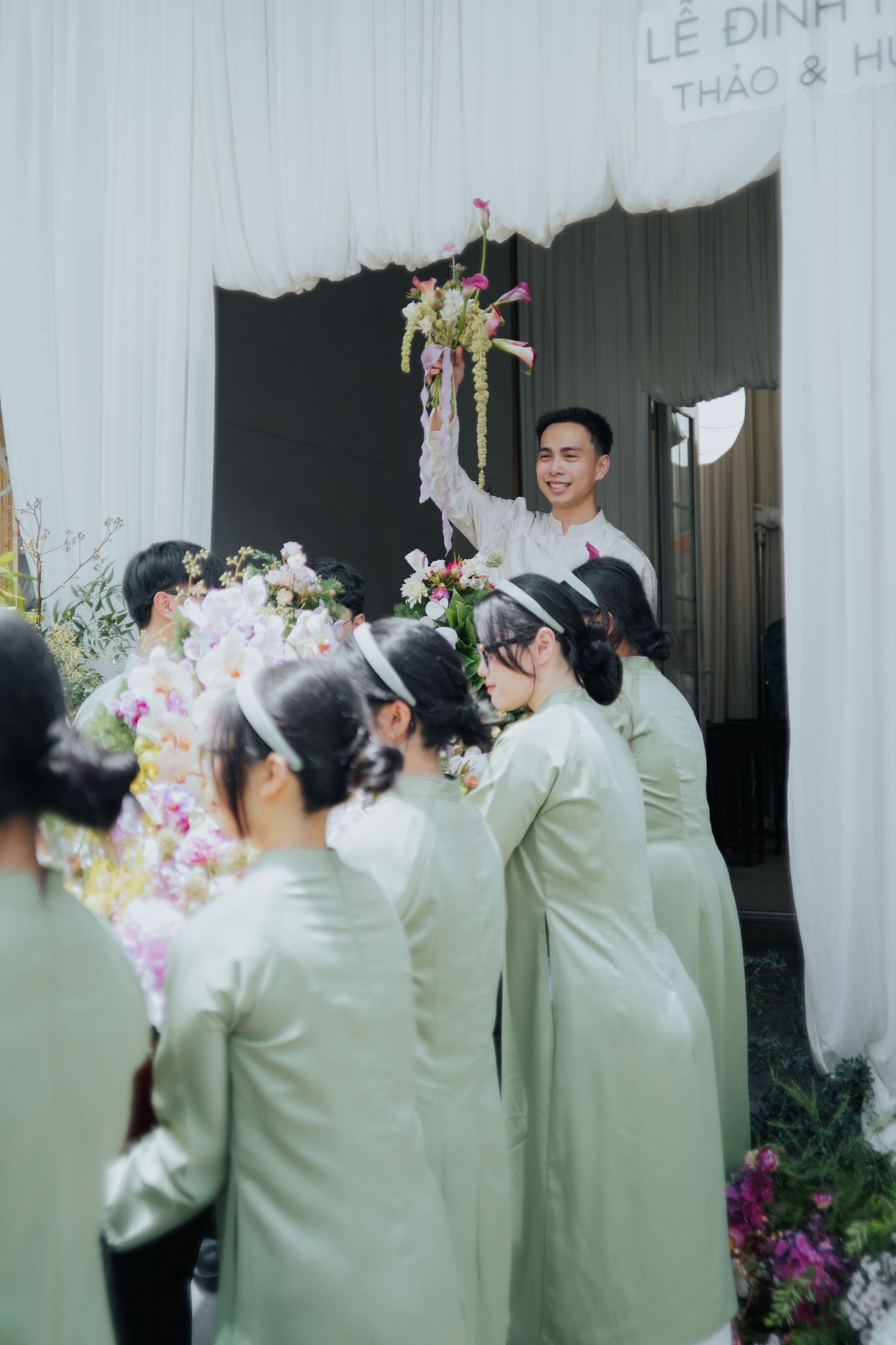 A smiling groom raises a floral bouquet at a Vietnamese wedding ceremony with bridesmaids in green ao dai.