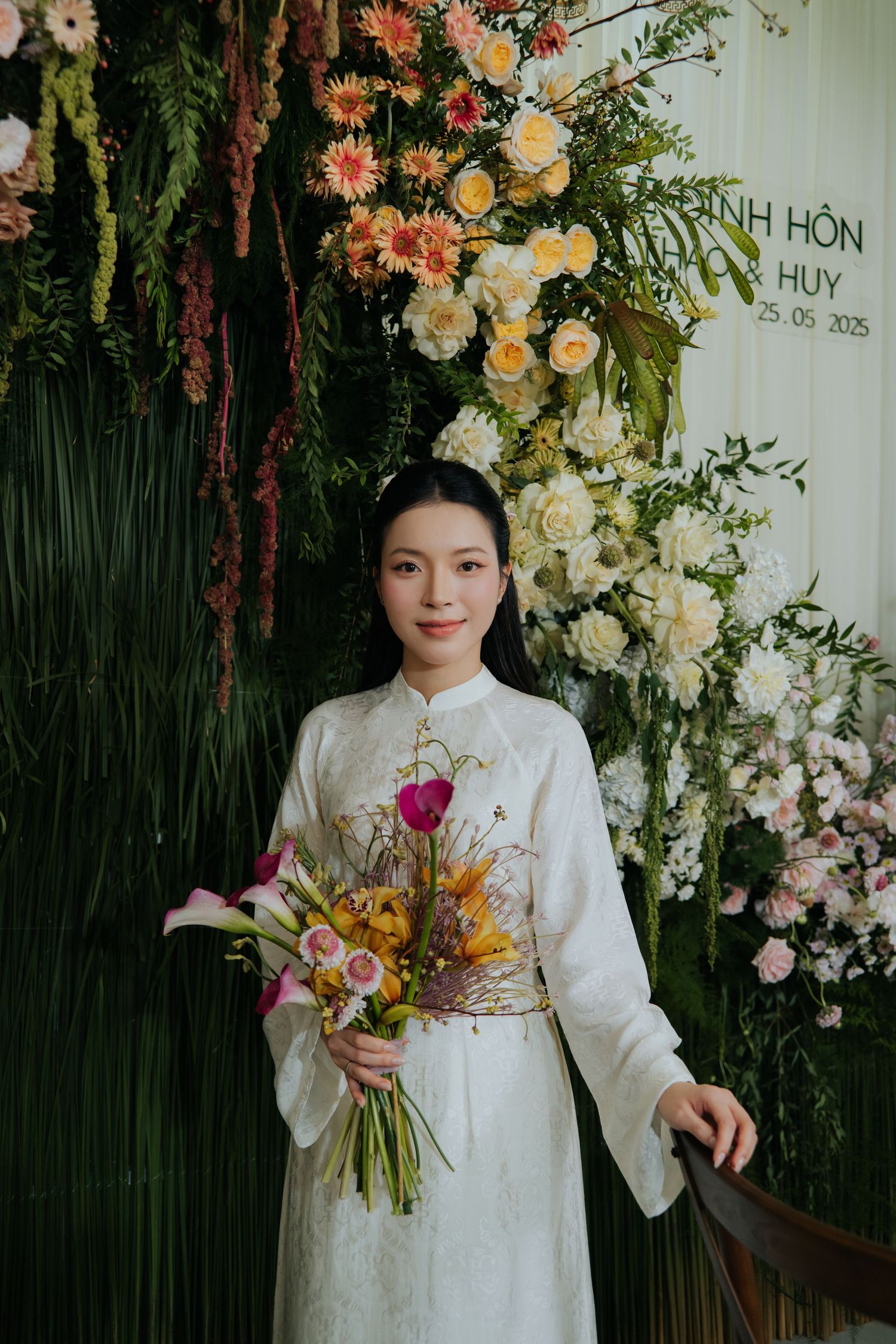 Vietnamese bride in white ao dai holds colorful bouquet, standing before elegant floral wedding backdrop dated 25.05.2025.