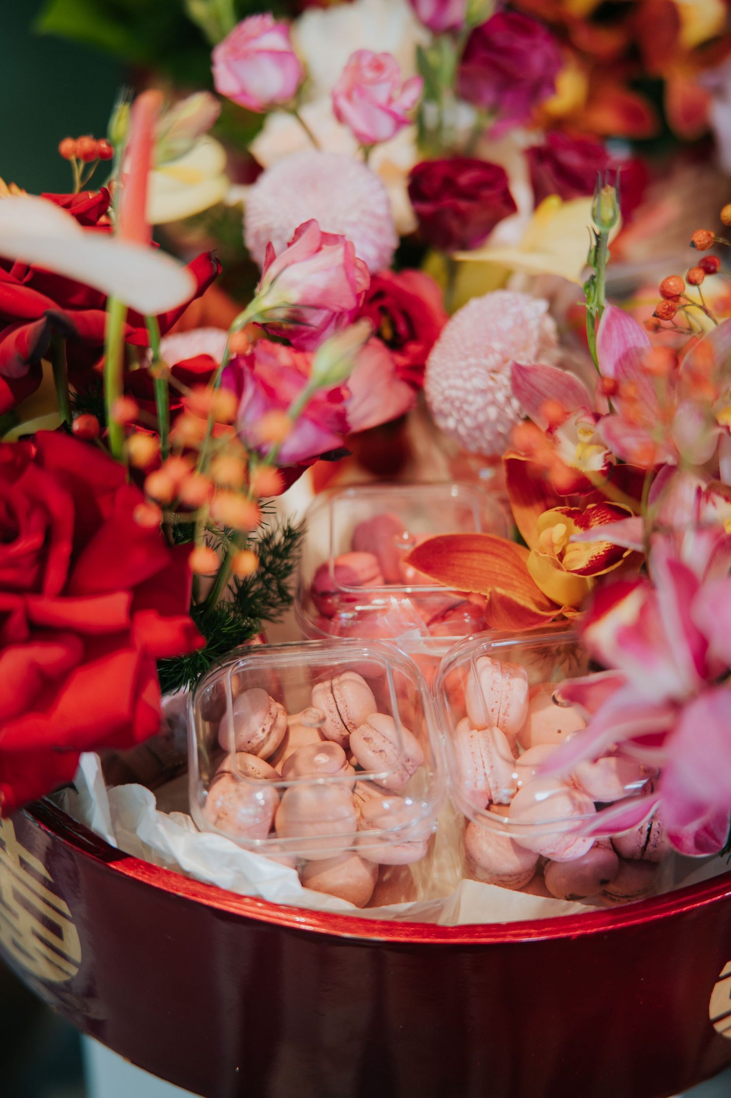 Pink macarons in clear packaging surrounded by vibrant red, pink, and orange floral arrangement in a red box.