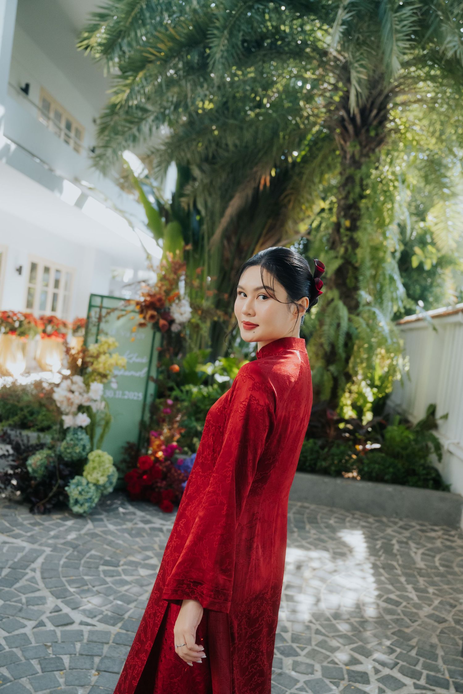 A young woman in a flowing red dress poses gracefully in a lush garden with tropical plants and colorful flowers.