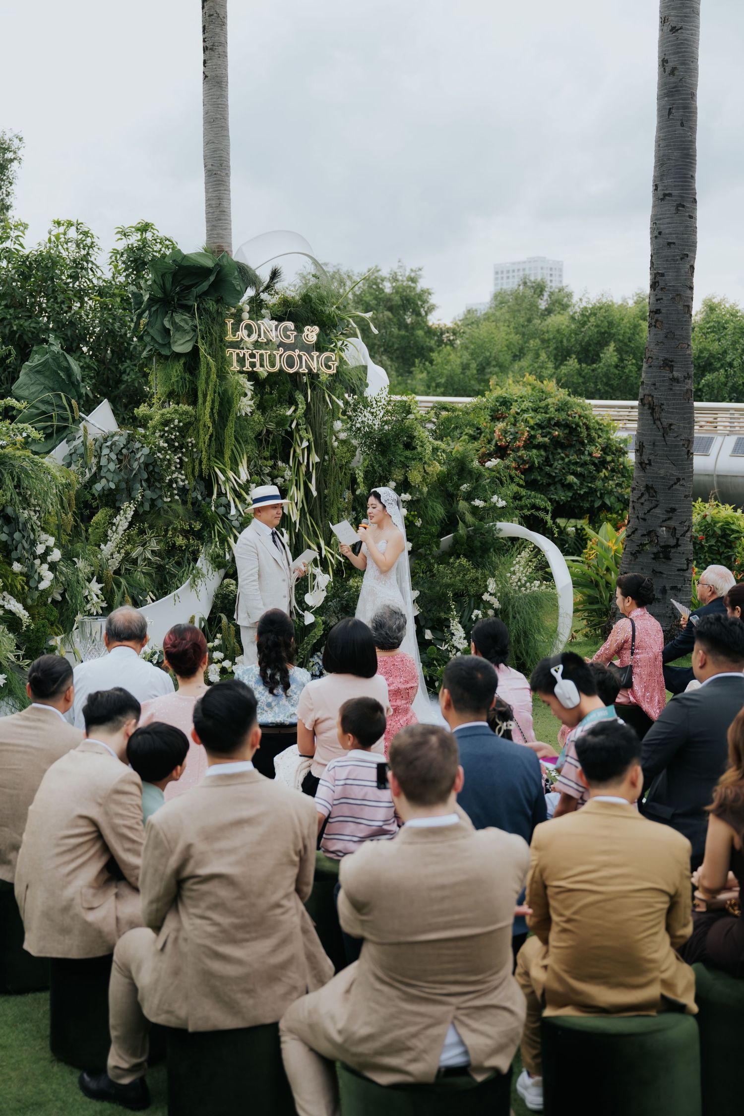 Outdoor garden wedding ceremony with guests seated as couple stands at floral arch reading Long & Thuong.