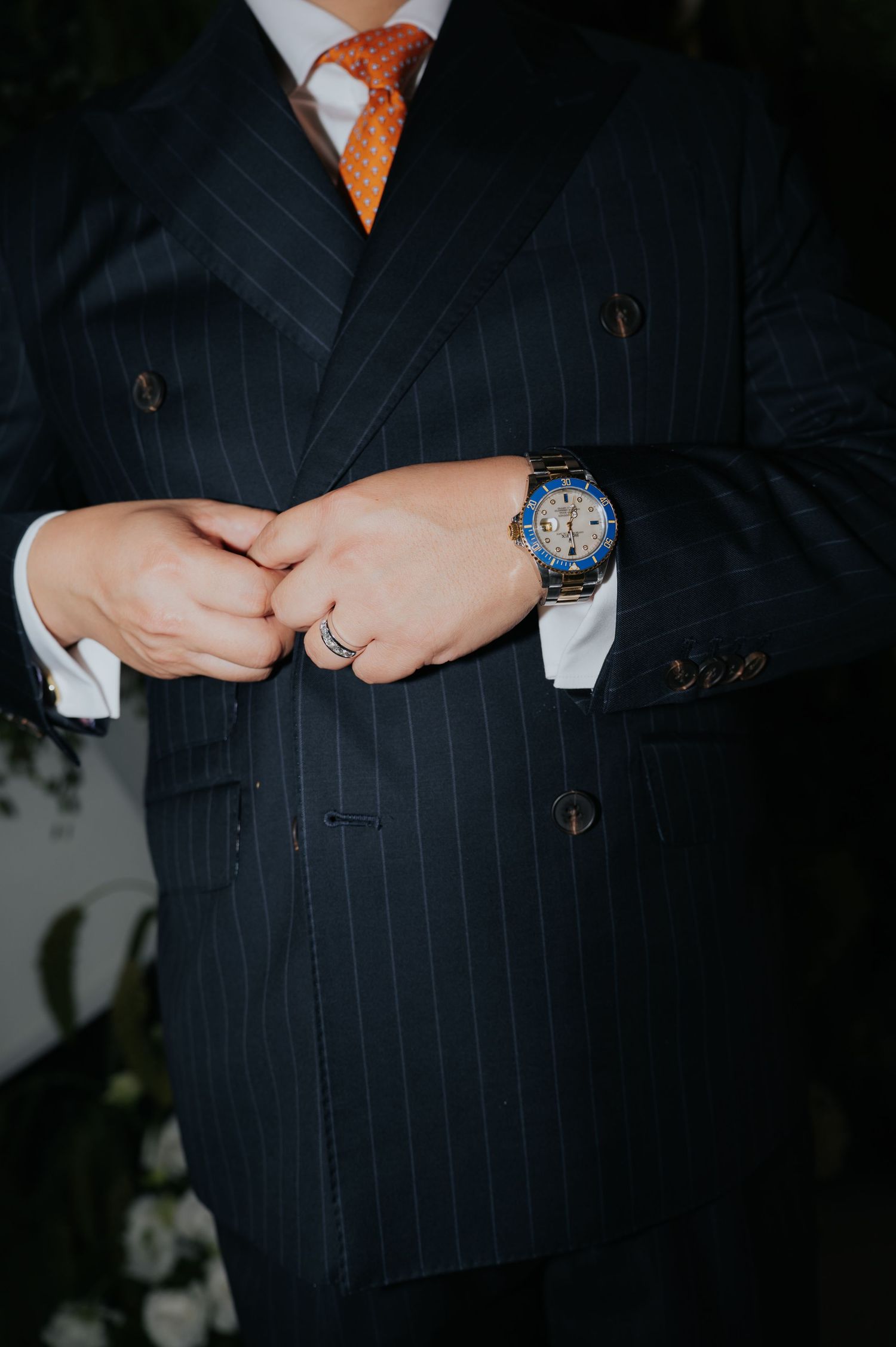 Man in navy pinstripe double-breasted suit with orange tie and blue watch adjusting jacket buttons.