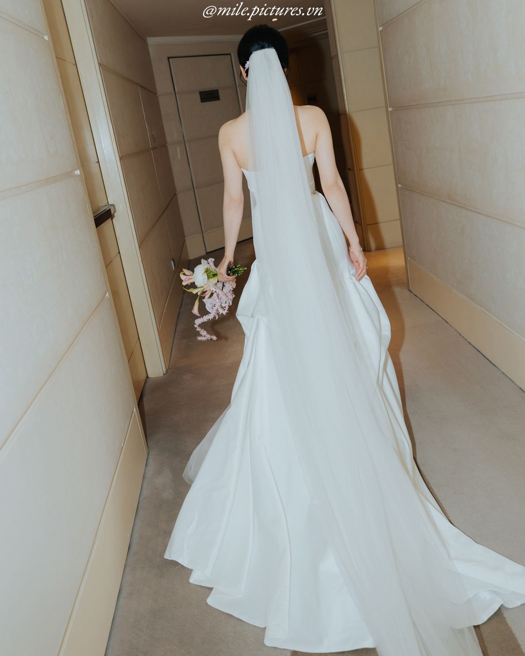 Bride in elegant white strapless wedding gown with long veil holding bouquet, viewed from behind in hallway.
