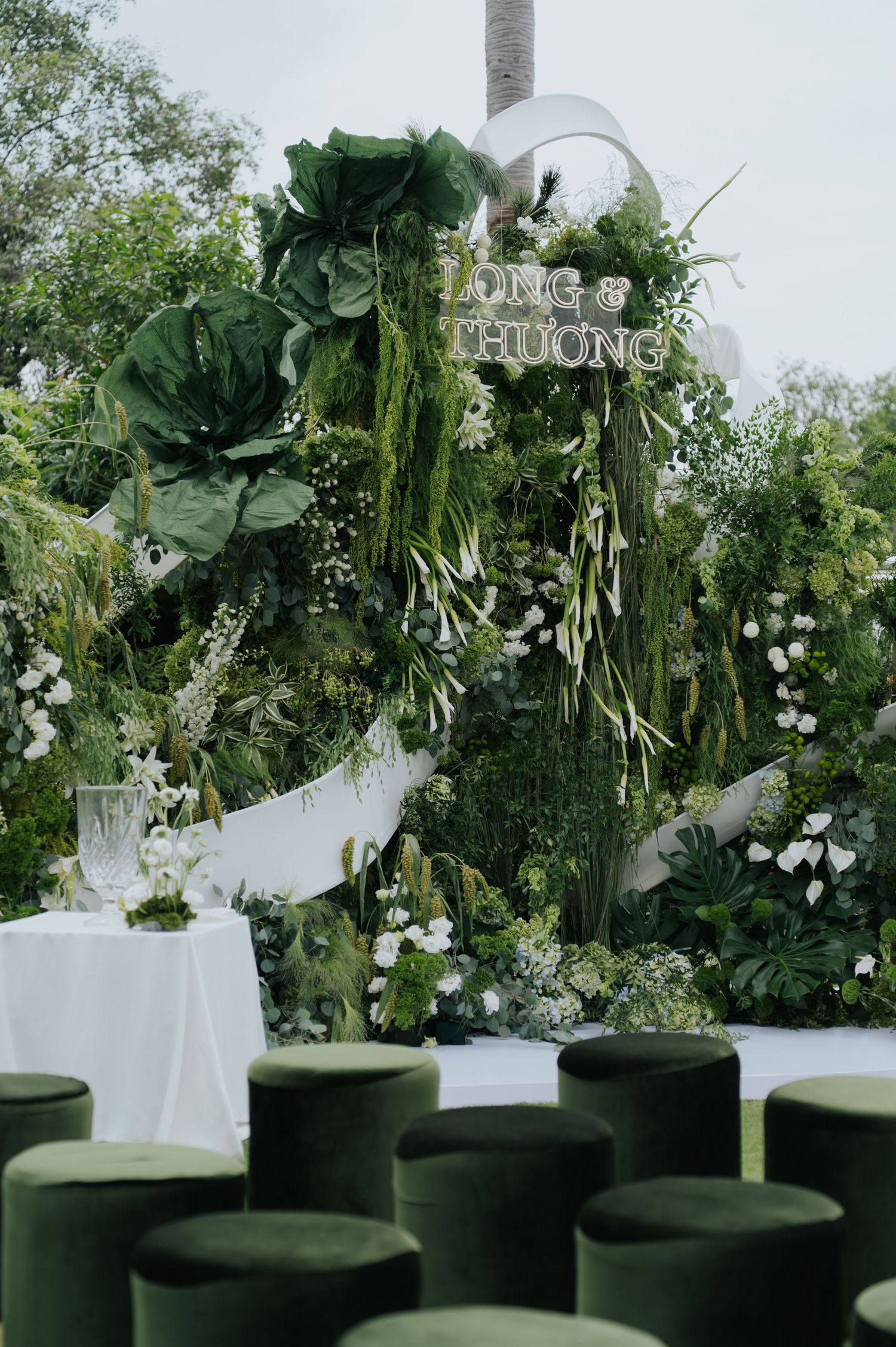 Lush green botanical wedding backdrop with white flowers, heart arch, and Long & Huong signage, surrounded by velvet ottomans.