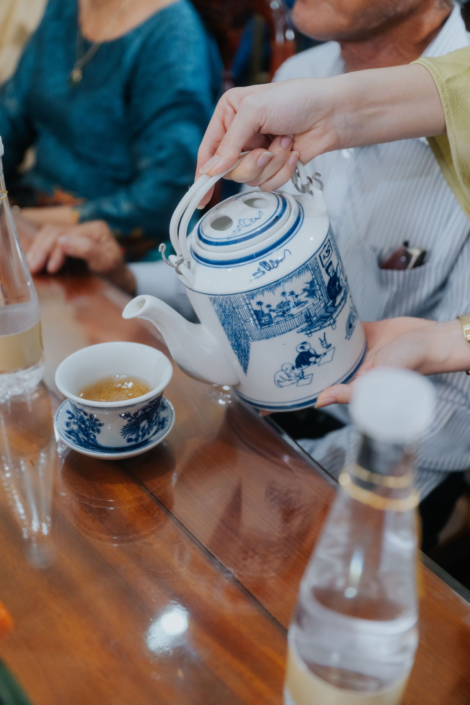 Person pouring tea from a blue and white Chinese porcelain teapot into a small cup on a wooden table.