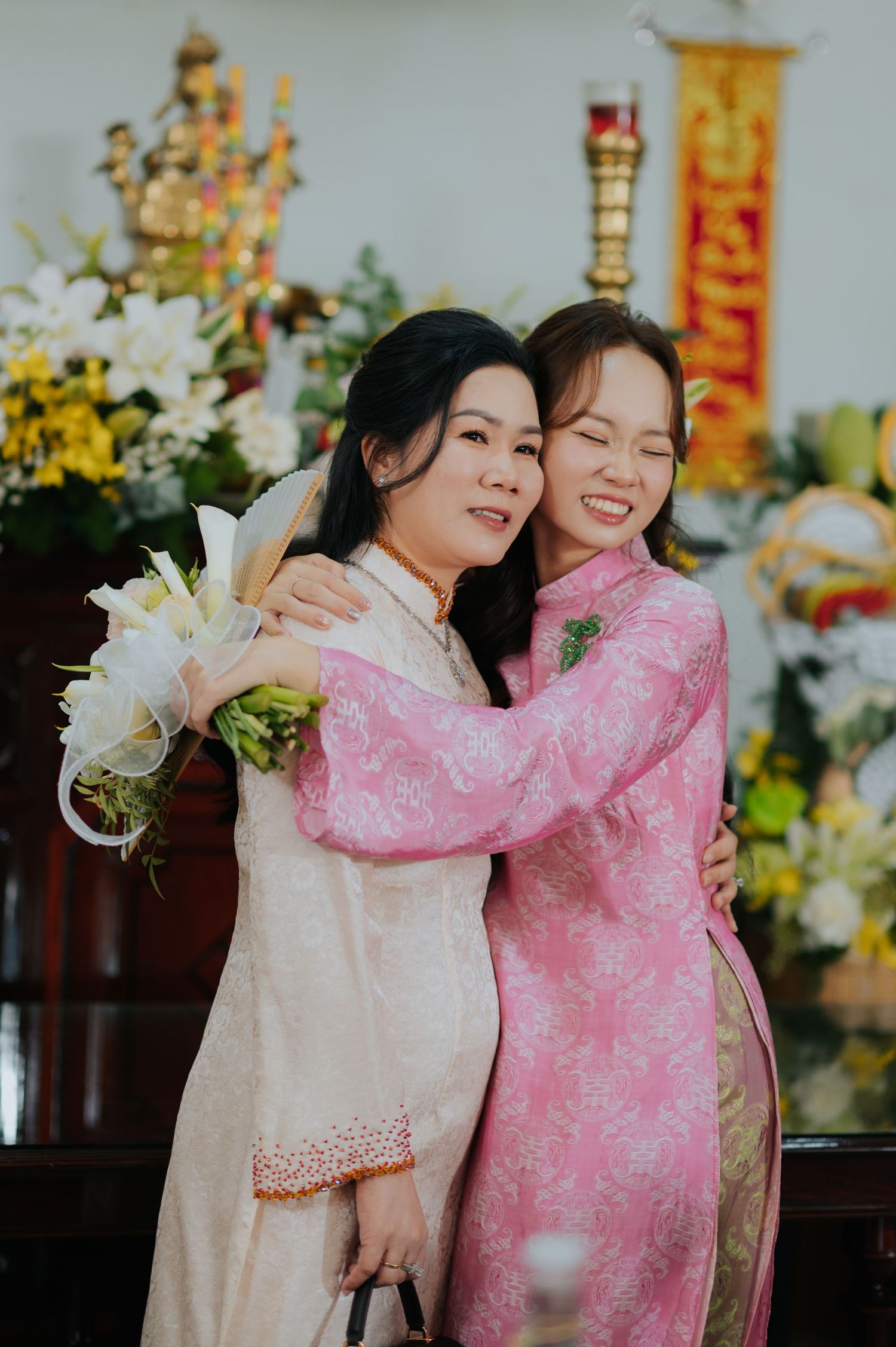 Two women in traditional Vietnamese ao dai embrace warmly, one in pink and one in cream, holding white flowers at a ceremony.
