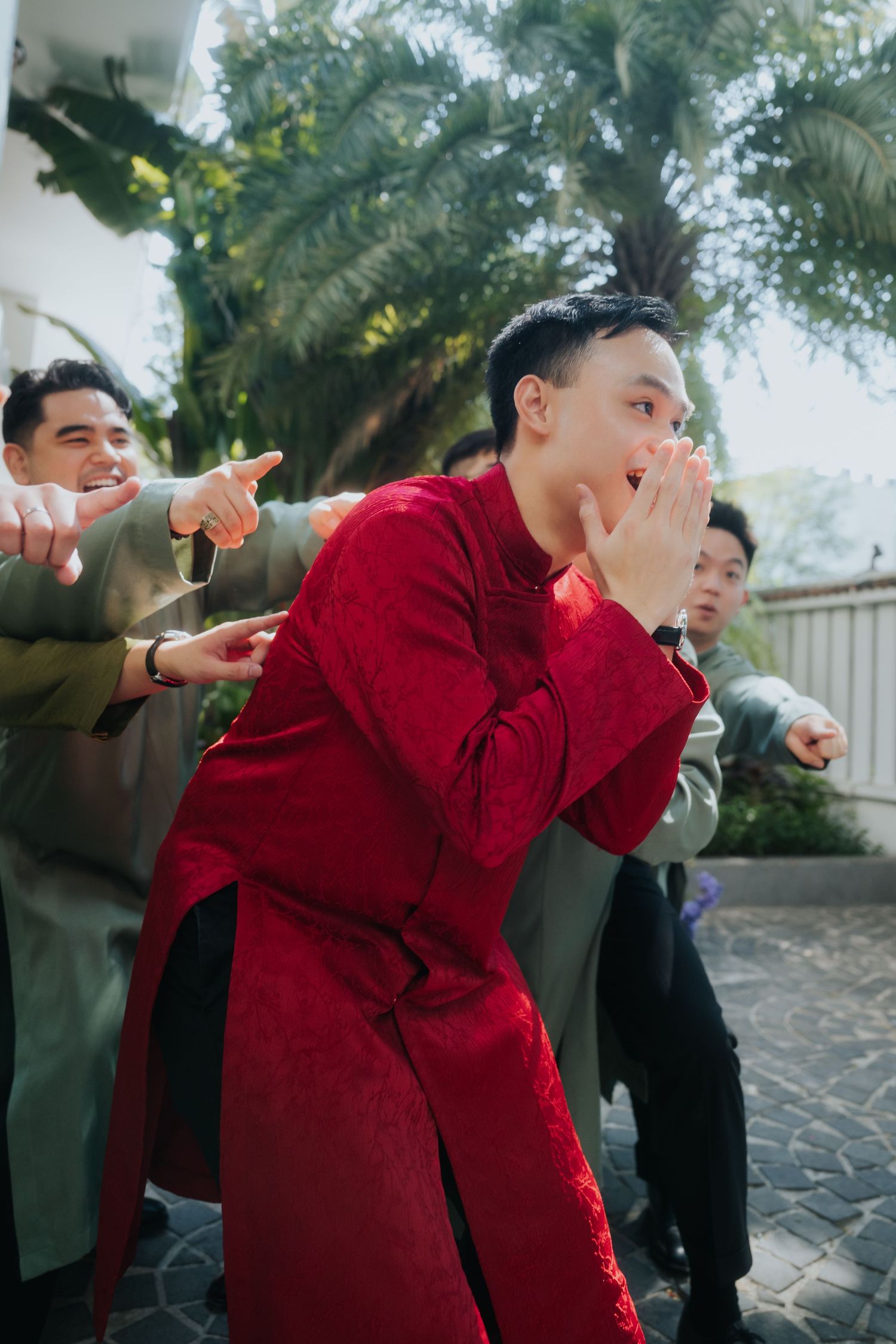 Young Asian man in red traditional ao dai reacts with surprise, flanked by friends pointing at him outdoors.
