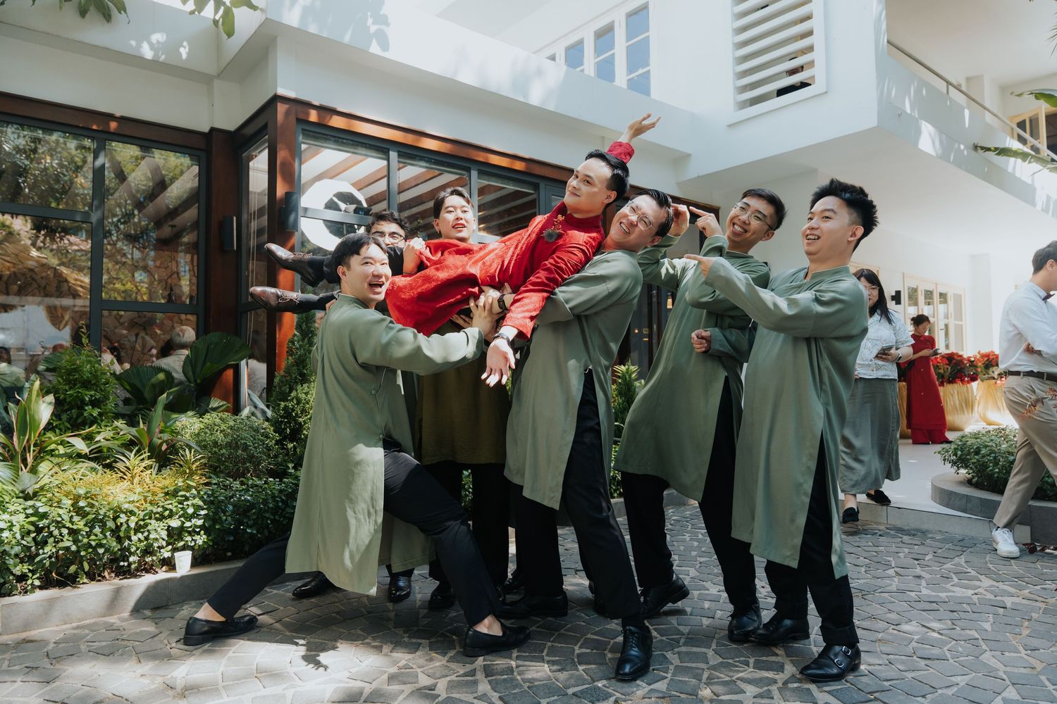 Group of people in traditional Vietnamese ao dai outfits joyfully lifting one person outdoors during a festive celebration.