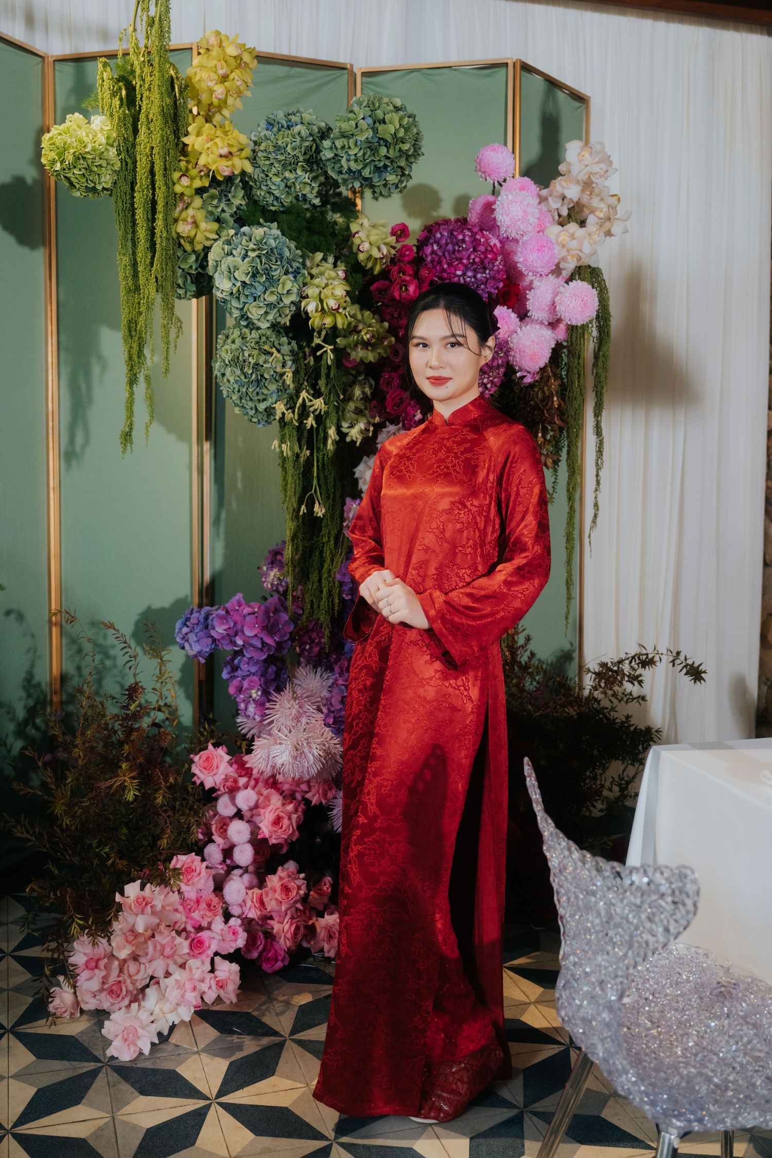 Woman in red ao dai gown poses before colorful floral backdrop with green folding screen at elegant event.