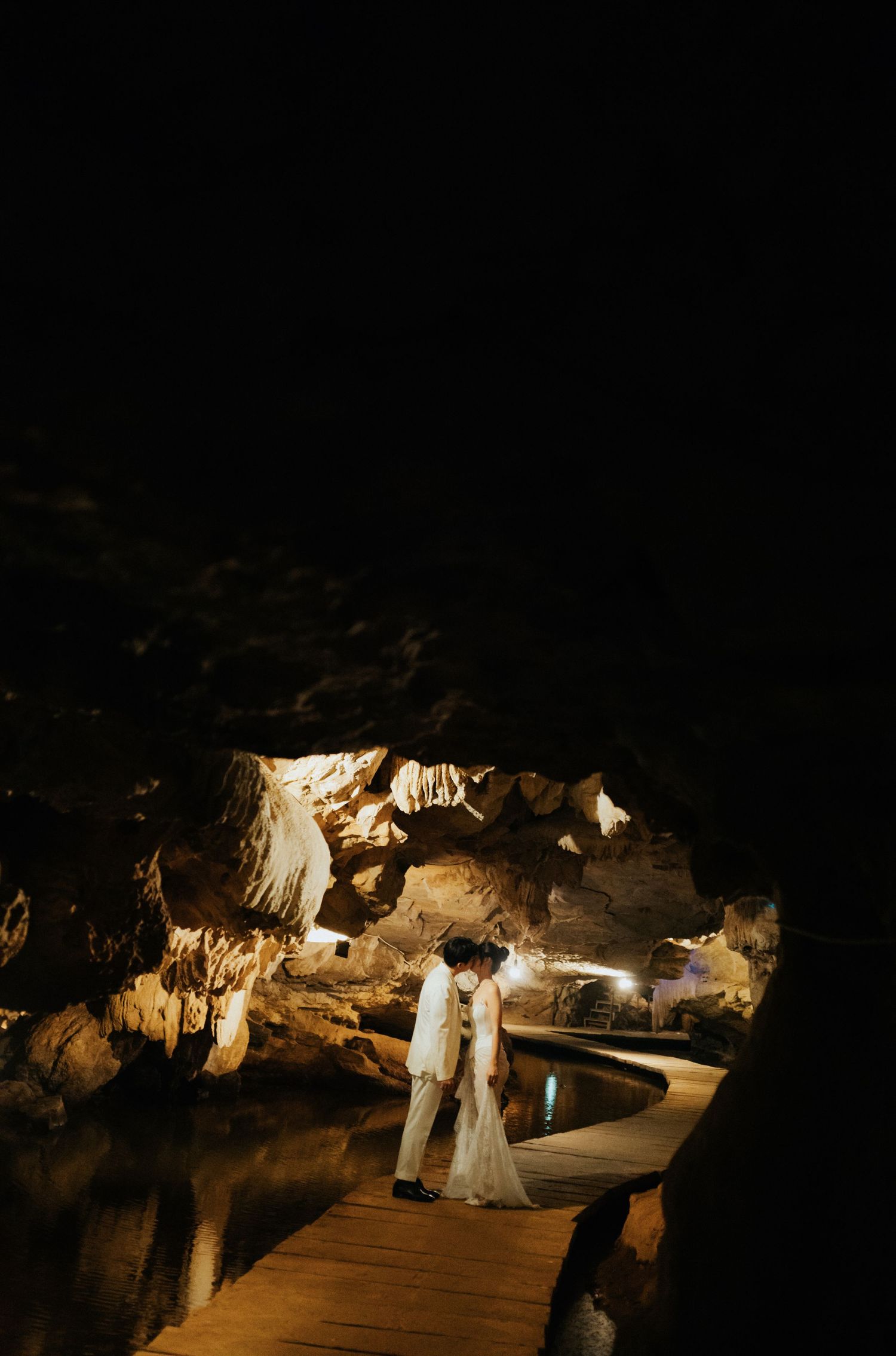 A couple shares a romantic moment on a wooden bridge inside a dramatic cave with glowing stalactites and a reflective pool.