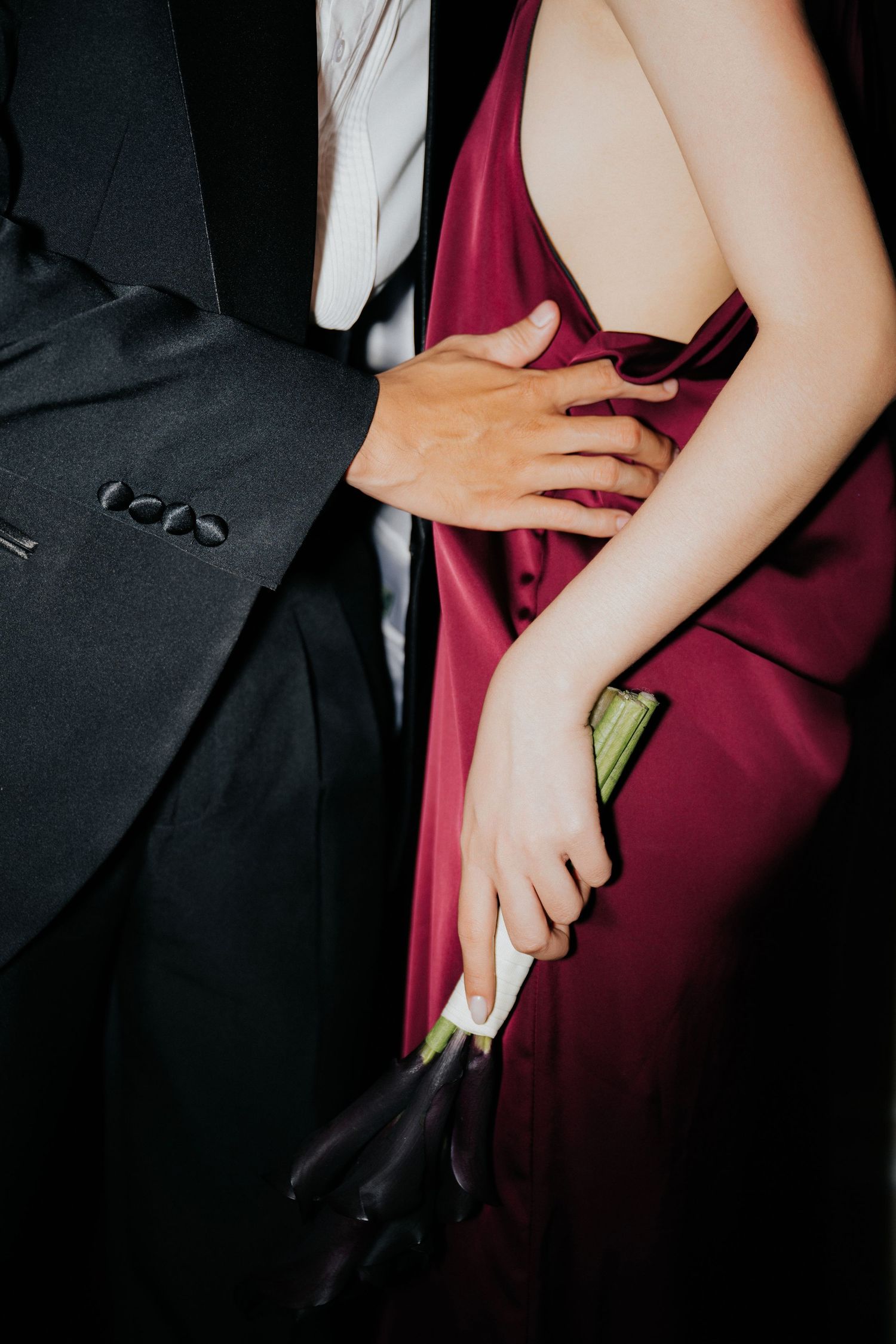 Couple in formal attire, man in black tuxedo placing hand on woman's back in burgundy gown holding dark flowers.