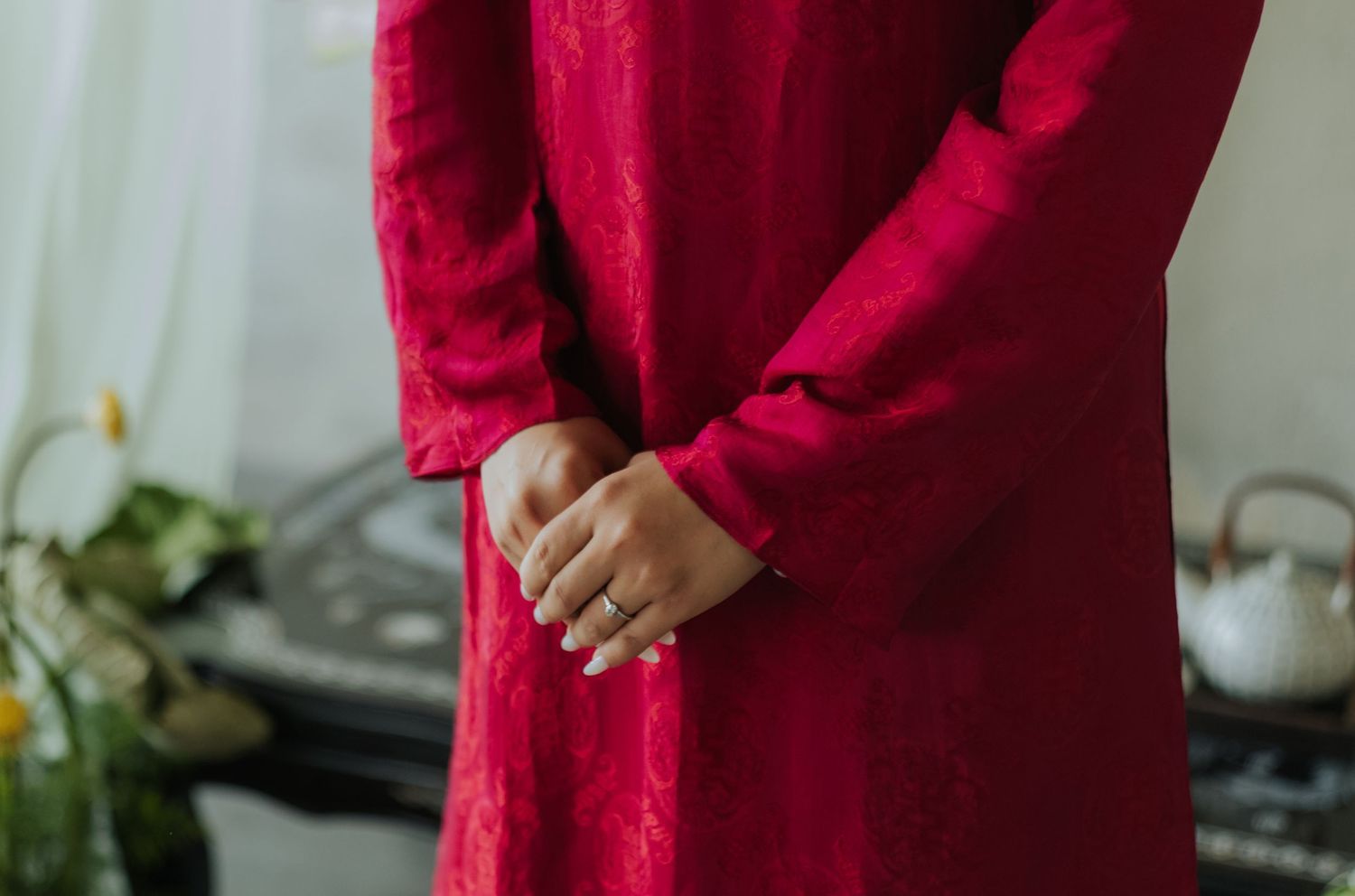 Woman in elegant red long-sleeve dress with clasped hands, wearing silver rings, standing in a softly lit room.